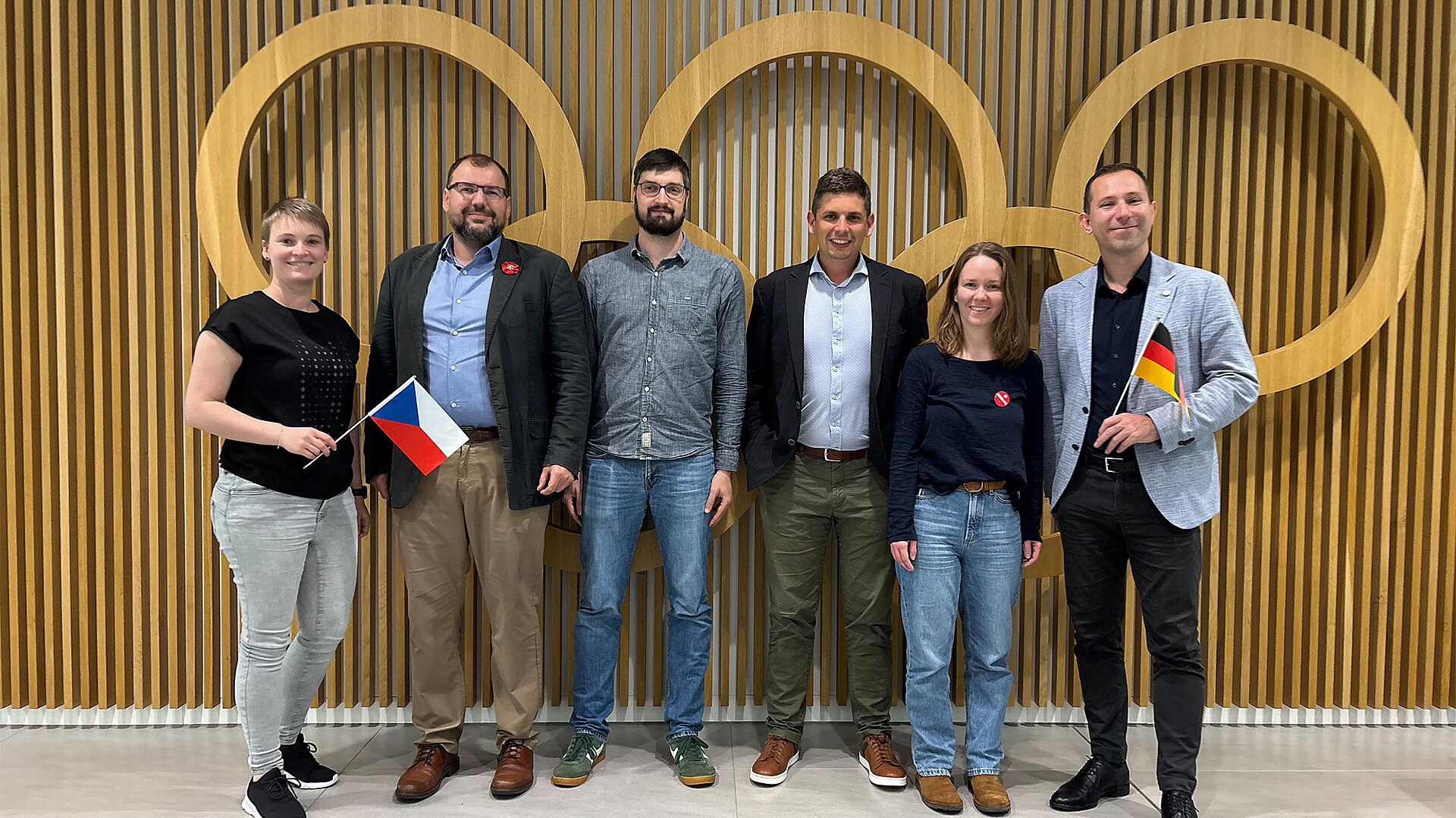 Sechs Personen stehen vor einer Wand mit olympischen Ringen, zwei halten die Flaggen von Tschechien und Deutschland, symbolisieren deutsch-tschechischen Jugendaustausch im Sport