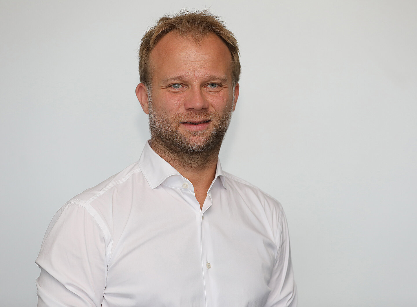 Ein Mann mit kurzem, blondem Haar und einem Bart steht vor einer weißen Wand. Er trägt ein weißes Hemd und lächelt leicht.