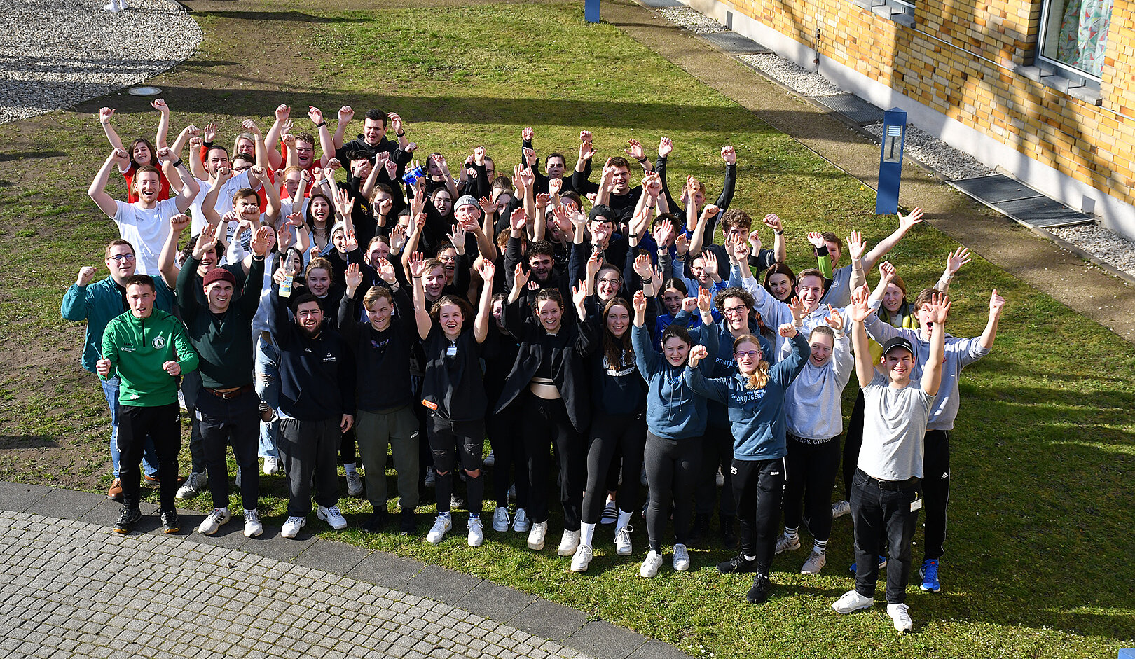 Eine große Gruppe von etwa 70 Personen steht im Freien und hebt die Arme in die Luft. Sie lächeln und scheinen sich zu freuen. Im Hintergrund sind Gebäude und ein grüner Rasen zu sehen.