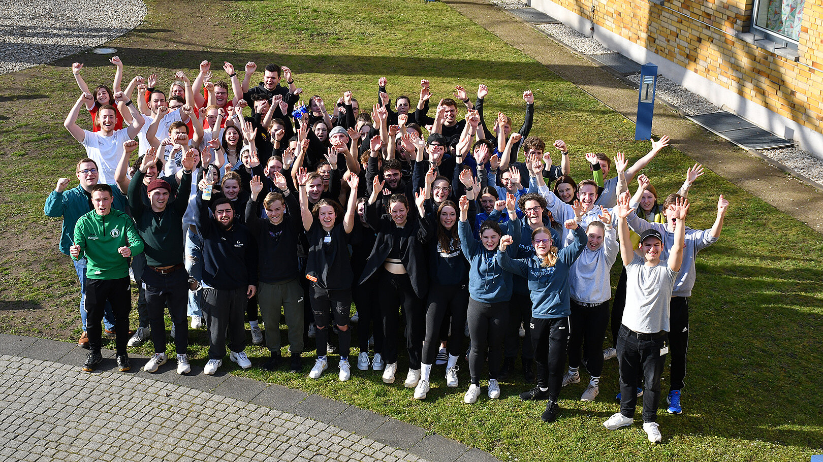 Eine große Gruppe von etwa 70 Personen steht im Freien und hebt die Arme in die Luft. Sie lächeln und scheinen sich zu freuen. Im Hintergrund sind Gebäude und ein grüner Rasen zu sehen.
