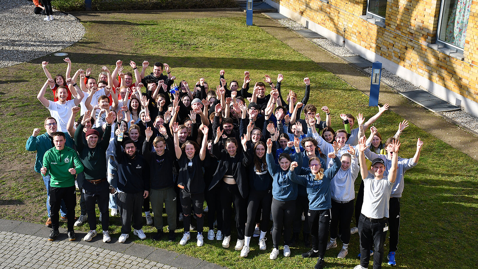 Eine große Gruppe von etwa 70 Personen steht im Freien und hebt die Arme in die Luft. Sie lächeln und scheinen sich zu freuen. Im Hintergrund sind Gebäude und ein grüner Rasen zu sehen.