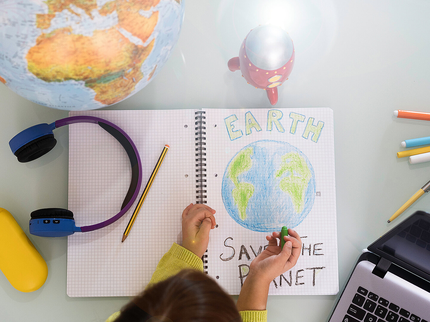 Ein Kind zeichnet in einem Notizbuch mit der Überschrift "EARTH" und dem Text "SAVE THE PLANET". Neben dem Notizbuch liegen Buntstifte, ein Laptop und ein Globus. Das Kind trägt Kopfhörer und hält einen grünen Stift.