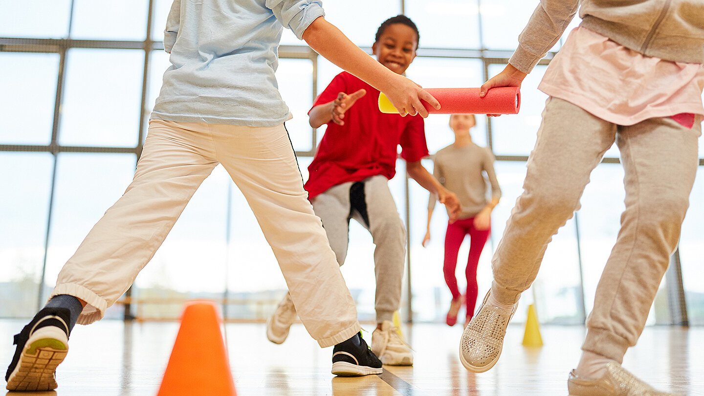 Eine Gruppe von Kindern spielt in einer Sporthalle. Sie übergeben einen Staffelstab und laufen um orangefarbene Kegel. Die Kinder tragen sportliche Kleidung und scheinen viel Spaß zu haben.