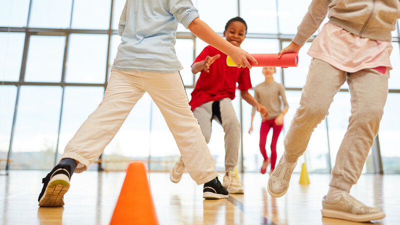 Kinder beim Staffellauf in der Turnhalle einer Schule, Übergabe eines roten Staffelstabs, orangefarbene Hütchen auf dem Boden