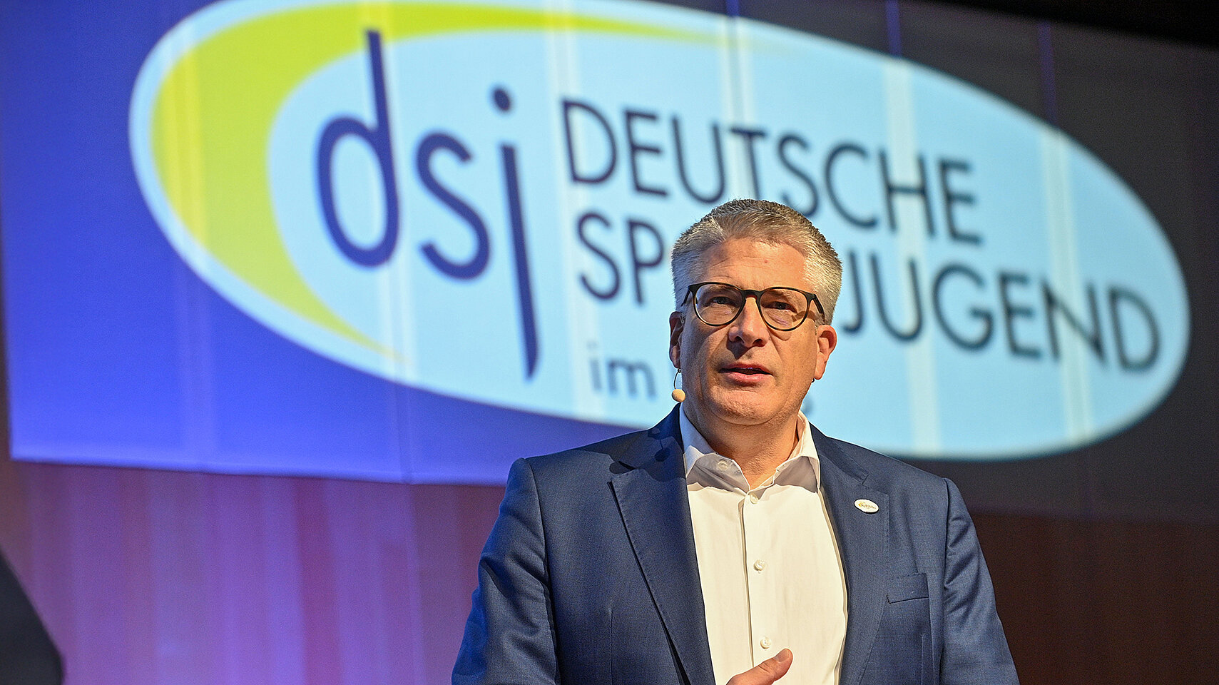Stefan Raid steht auf einer Bühne vor einer beleuchteten Leinwand mit dem Logo der Deutschen Sportjugend (dsj) und hält eine Präsentation.