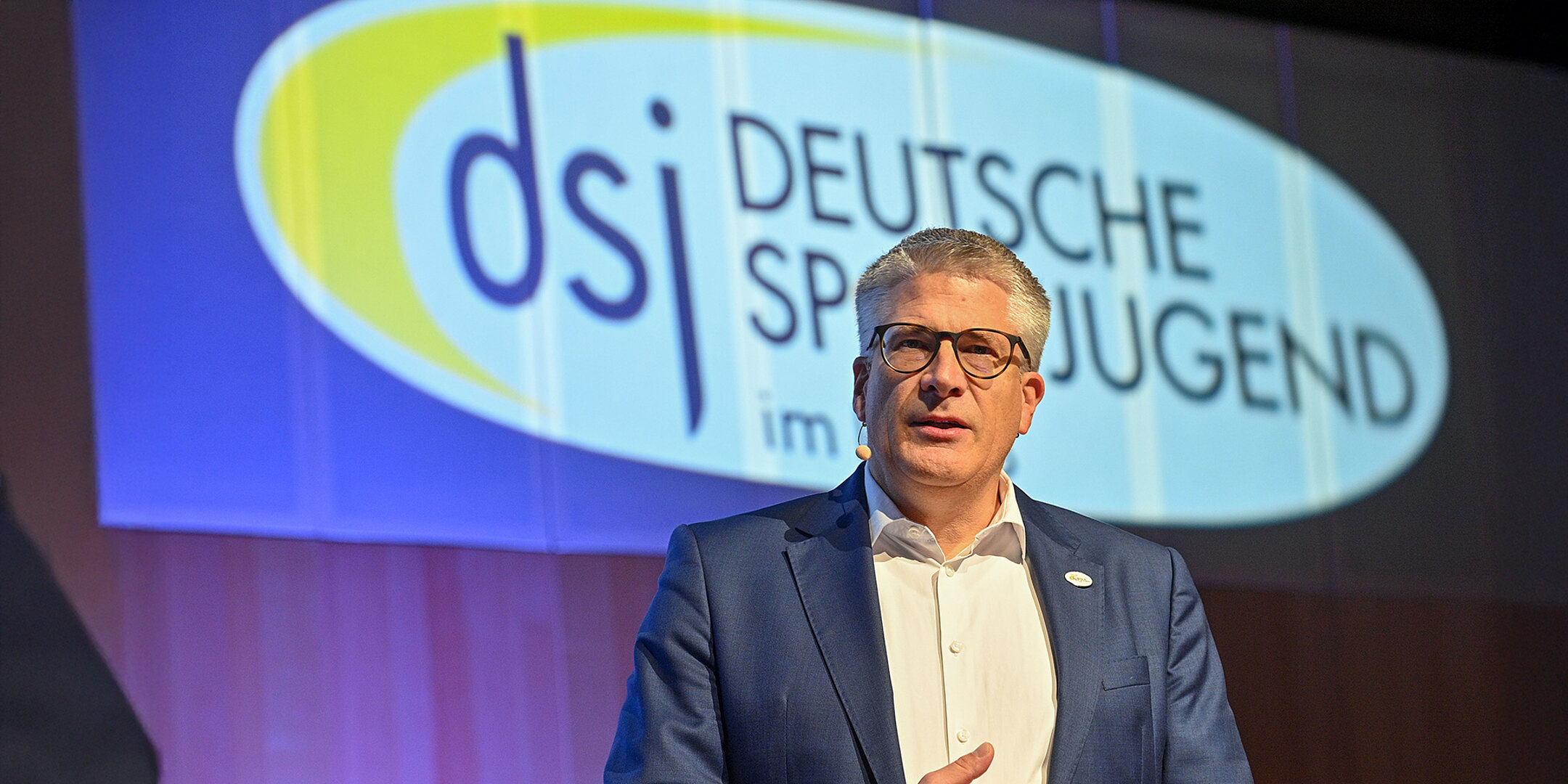 Stefan Raid, Vorsitzender der Deutschen Sportjugend (dsj), spricht auf einer Bühne vor dem projizierten dsj-Logo.
