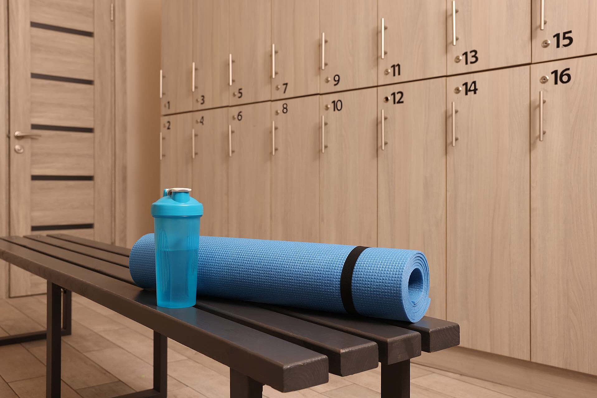 Eine blaue Yogamatte liegt auf einer Bank in einem Umkleideraum. Daneben steht eine transparente Trinkflasche in Blau. Im Hintergrund sind Holzschränke mit nummerierten Schlössern zu sehen.