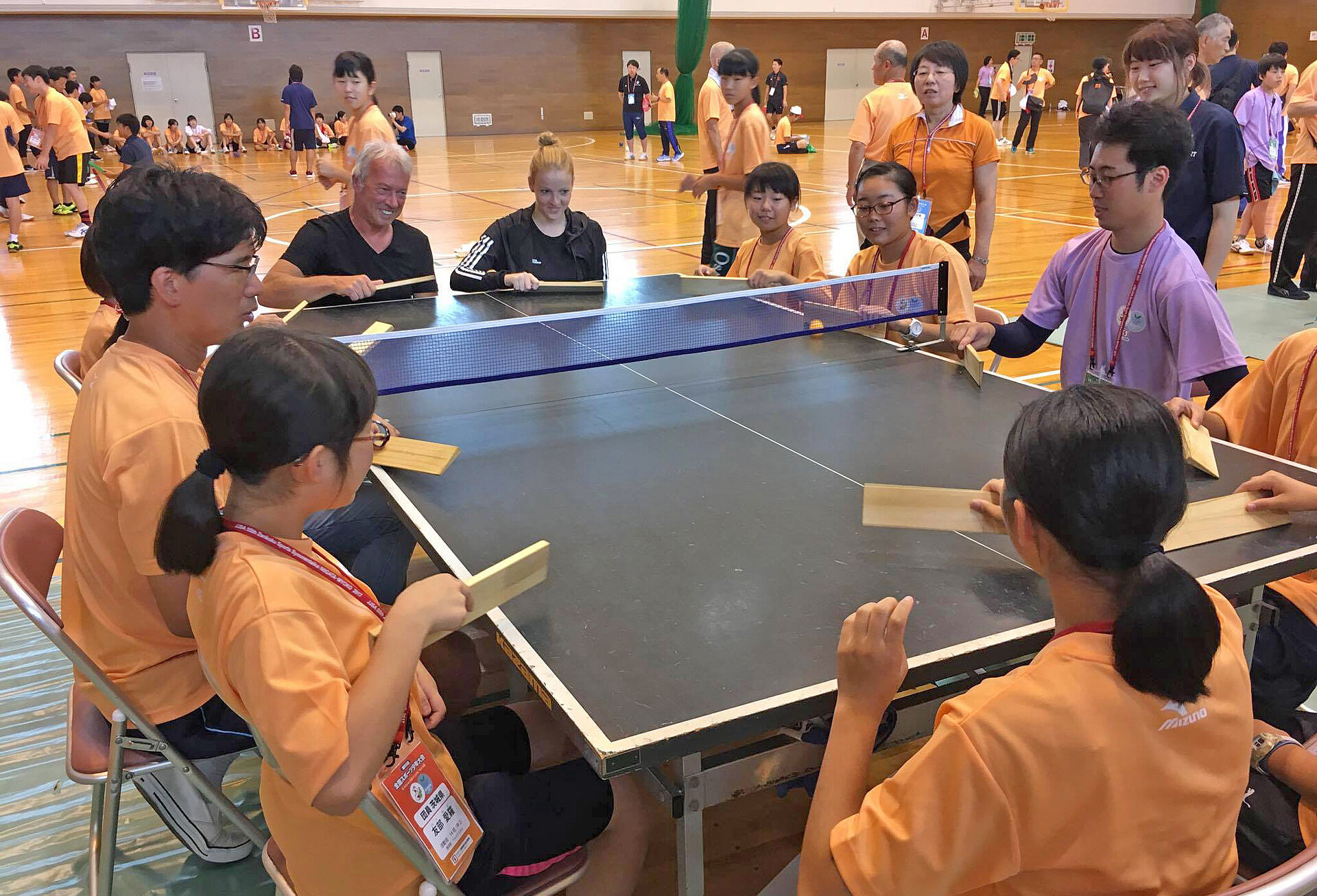 Eine Gruppe von Kindern und Erwachsenen spielen an einer Tischtennisplatte ein Spiel in einer Sporthalle. Alle tragen orangefarbene T-Shirts. Einige sitzen an einem Tisch, während andere stehen und zuschauen. Die Atmosphäre ist freundlich und gemeinschaftlich.