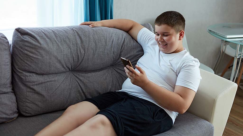 ein übergewichtiger Junge liegt auf der Couch und schaut in sein Handy