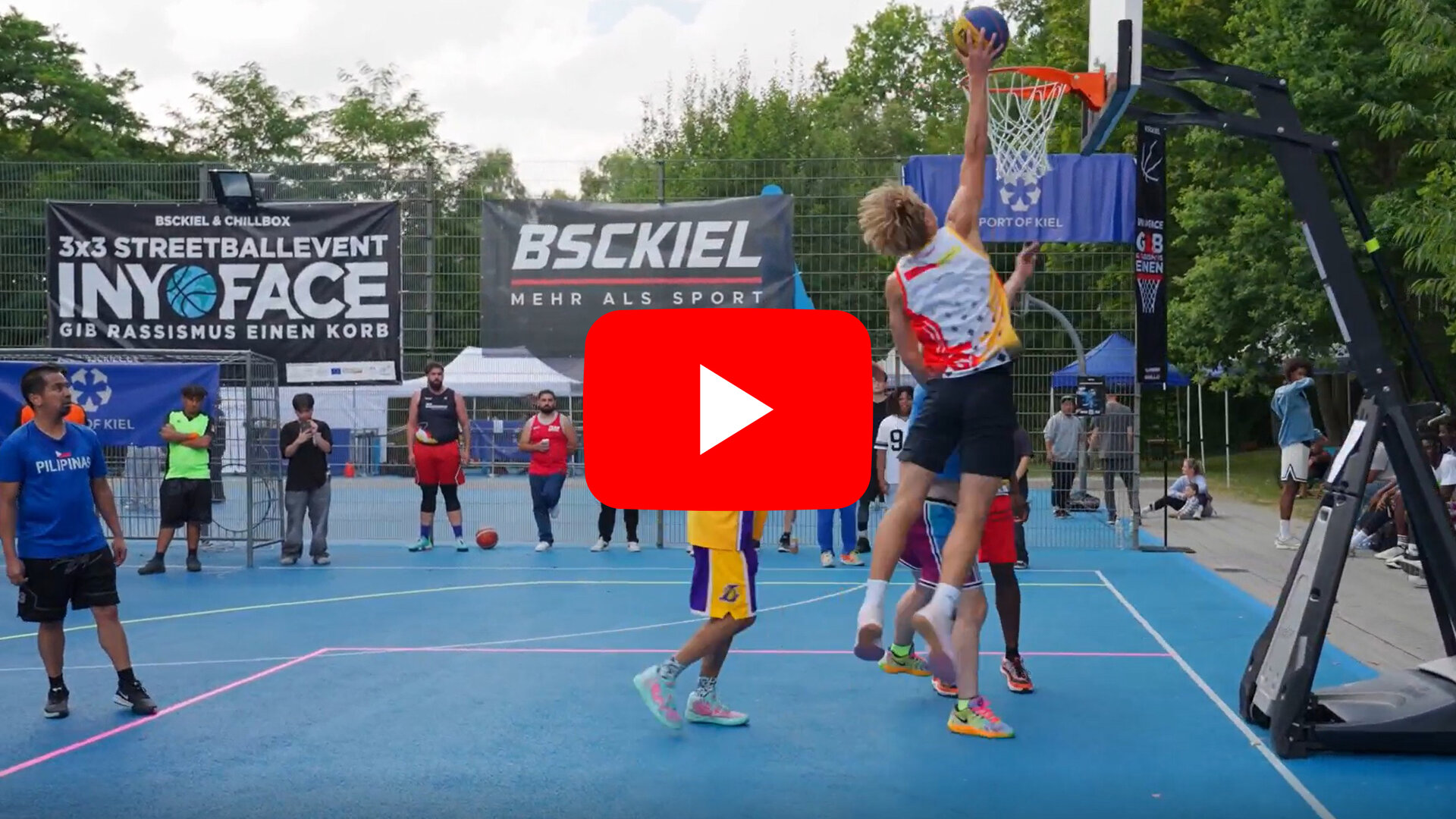 Auf einem blauen Basketballfeld spielen mehrere Jugendliche Basketball. Ein Spieler in einem weißen Trikot mit roten und gelben Akzenten springt hoch und versucht, den Ball in den Korb zu werfen. Um ihn herum stehen weitere Spieler in bunten Sportkleidungen, die das Spiel verfolgen oder verteidigen. Im Hintergrund sind zwei große Banner zu sehen: Einer trägt die Aufschrift '3x3 STREETBALLEVENT INY FACE GIB RASSISMUS EINEN KORB', der andere zeigt das Logo 'BSCKIEL MEHR ALS SPORT'. Bäume und ein Zaun umgeben das Spielfeld.