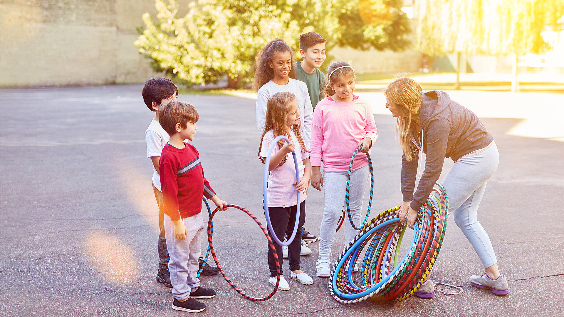 Eine erwachsene Frau beugt sich nach vorne und hält mehrere bunte Hula-Hoop-Reifen in der Hand. Vor ihr stehen fünf Kinder in einer Reihe, die jeweils einen Hula-Hoop-Reifen halten. Die Kinder tragen legere Kleidung in verschiedenen Farben, darunter Rot, Weiß, Rosa und Hellblau. Die Szene spielt sich im Freien ab, im Hintergrund sind unscharfe grüne Büsche und eine Mauer zu sehen. Die Lichtverhältnisse sind hell und sonnig.