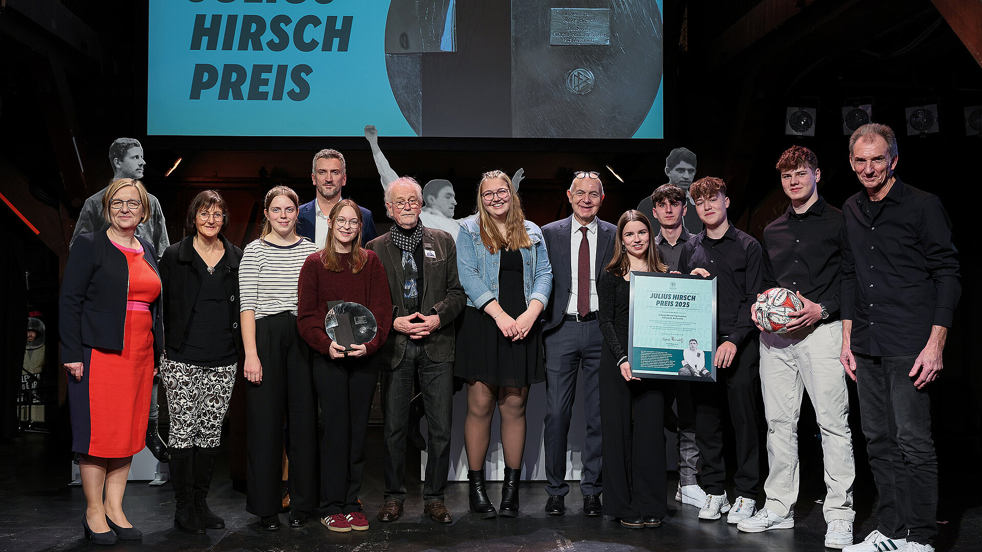 Eine Gruppe von Menschen steht auf einer Bühne vor einer großen Leinwand mit der Aufschrift 'JULIUS HIRSCH PREIS'. Einige Personen halten Auszeichnungen: eine Person hält eine runde Trophäe, eine andere eine Urkunde, und eine weitere Person hält einen Fußball. Die Personen tragen unterschiedliche Kleidung, darunter ein rotes Kleid, ein gestreiftes Oberteil, ein Anzug mit Krawatte und legere Kleidung. Die Bühne ist dunkel, und im Hintergrund sind weitere Auszeichnungen sichtbar.