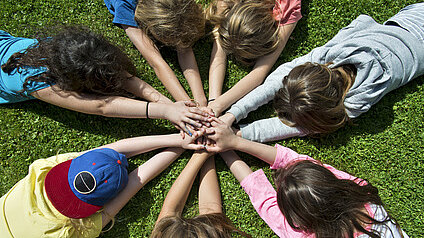 Eine Gruppe von Kindern liegt im Kreis auf dem Gras und legt ihre Hände übereinander in die Mitte. Die Kinder tragen bunte T-Shirts und zeigen eine gemeinschaftliche Geste.