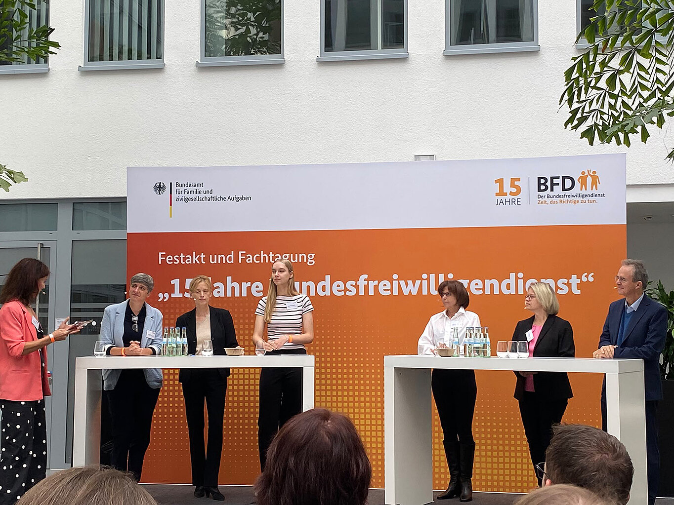 Eine Veranstaltung mit mehreren Personen an Stehtischen, die auf einer Bühne stehen. Im Hintergrund ein Banner mit dem Text „15 Jahre BFD“ und „Festakt und Fachtagung“. Die Anwesenden diskutieren, während eine Person spricht.