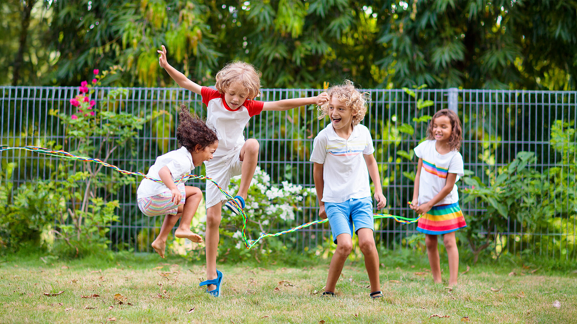Vier Kinder spielen im Garten mit einem Springseil, zwei springen, zwei halten das Seil, um den 'Day of Play' am 11. Juni zu feiern. Vom Weltspieltag zum Day of Play.