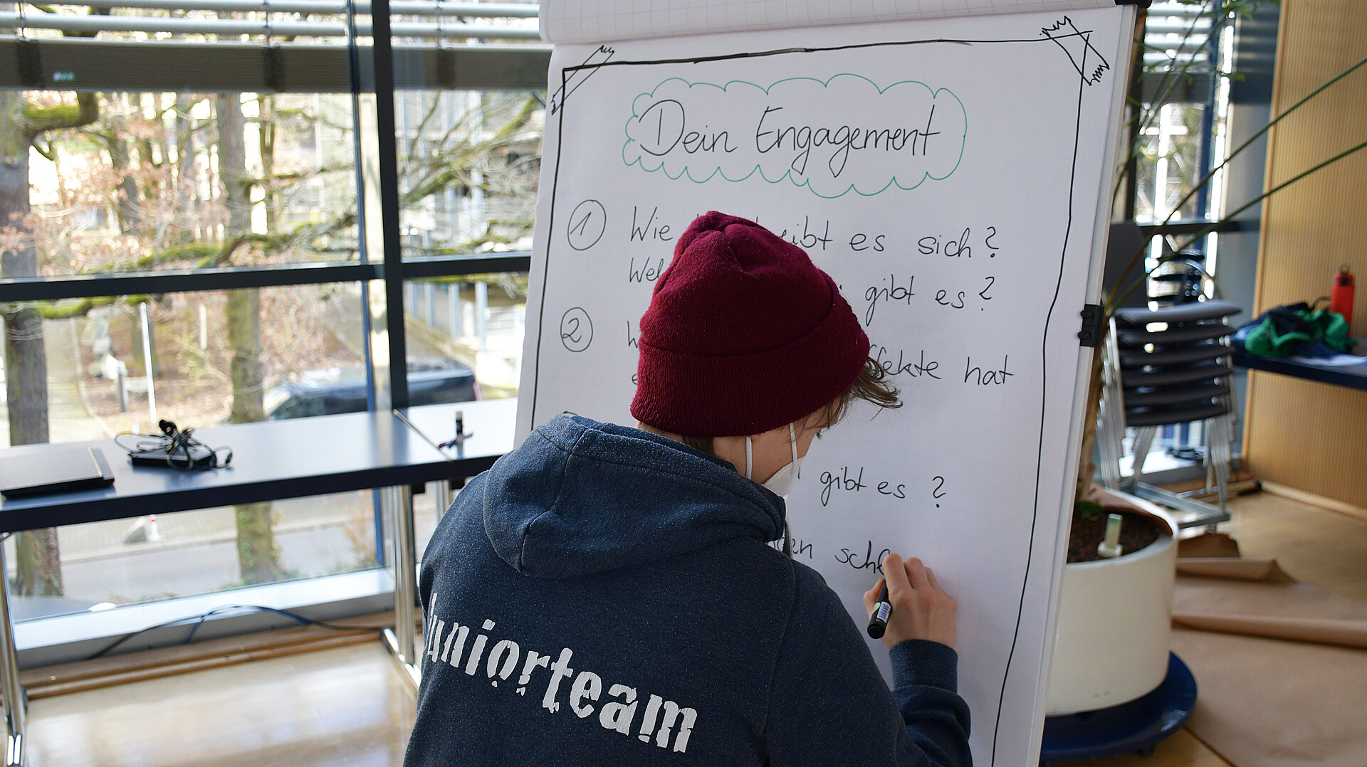 Person mit roter Mütze und blauem Pullover mit Aufschrift 'Juniorteam' schreibt auf ein Flipchart mit der Überschrift 'Dein Engagement' und mehreren Fragen