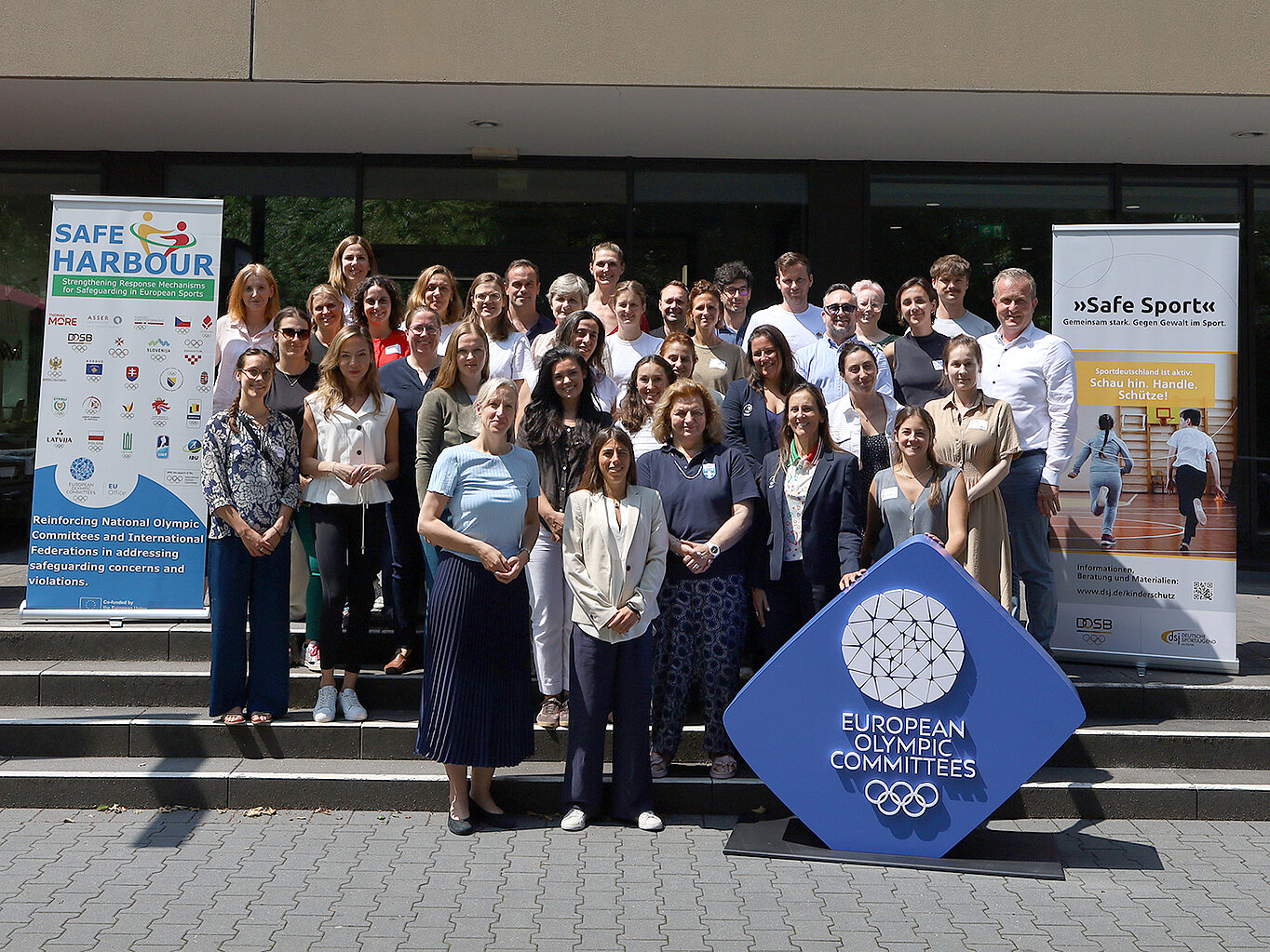 Eine Gruppe von etwa 40 Personen steht vor einem Gebäude, umgeben von Bannern, die mit olympischen Themen und Sportorganisationen zu tun haben. Im Vordergrund befindet sich ein großes, blaues Logo der Europäischen Olympischen Komitees. Die Teilnehmer lächeln und posieren für das Foto.