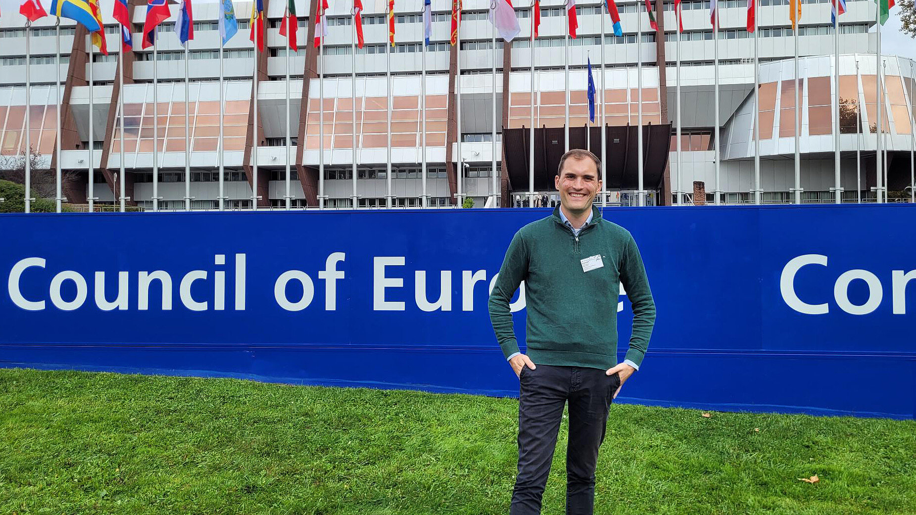 Ein Mann, Luca Wernert Vice-Chair von ENGSO Youth und dsj-Vorstandsmitglied steht auf einer Rasenfläche vor einem blauen Banner mit der weißen Aufschrift 'Council of Europe'. Er trägt einen grünen Pullover über einem hellblauen Hemd, eine schwarze Hose und hat die Hände in den Hosentaschen. Im Hintergrund sind zahlreiche Flaggen verschiedener Länder vor einem mehrstöckigen Gebäude zu sehen.