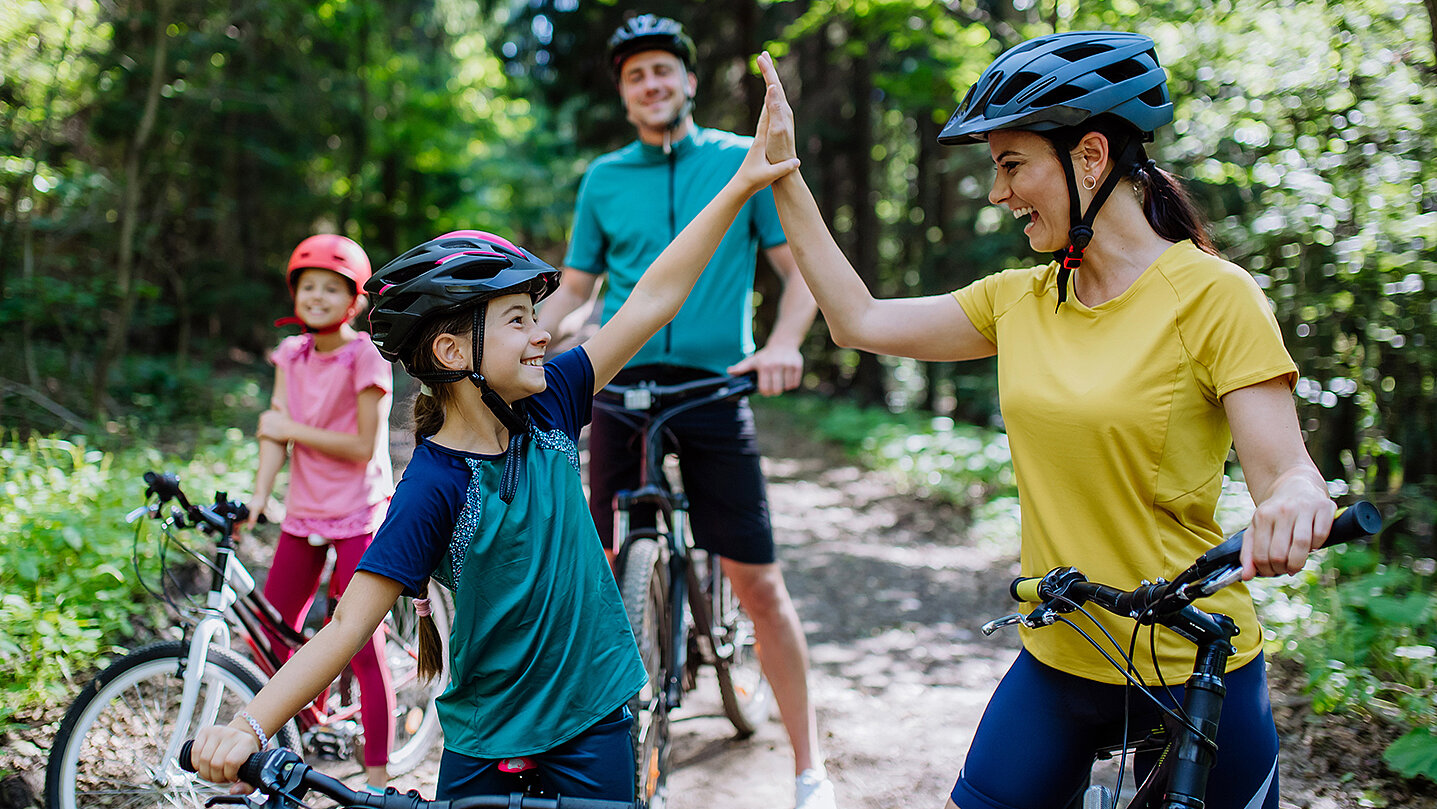 Eine Frau und ein Mädchen in Fahrradhelmen geben sich im Wald ein High-Five. Im Hintergrund stehen zwei weitere Personen auf Fahrrädern. Die Szene strahlt Freude und Zusammenhalt aus.