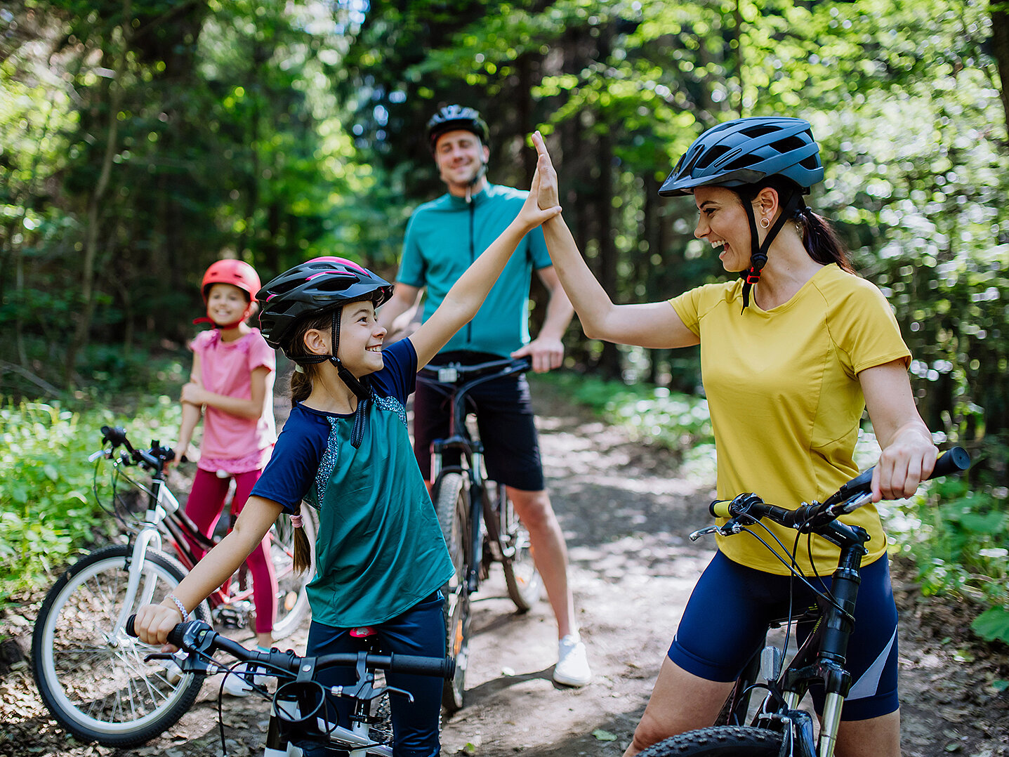 Eine Frau und ein Mädchen in Fahrradhelmen geben sich im Wald ein High-Five. Im Hintergrund stehen zwei weitere Personen auf Fahrrädern. Die Szene strahlt Freude und Zusammenhalt aus.