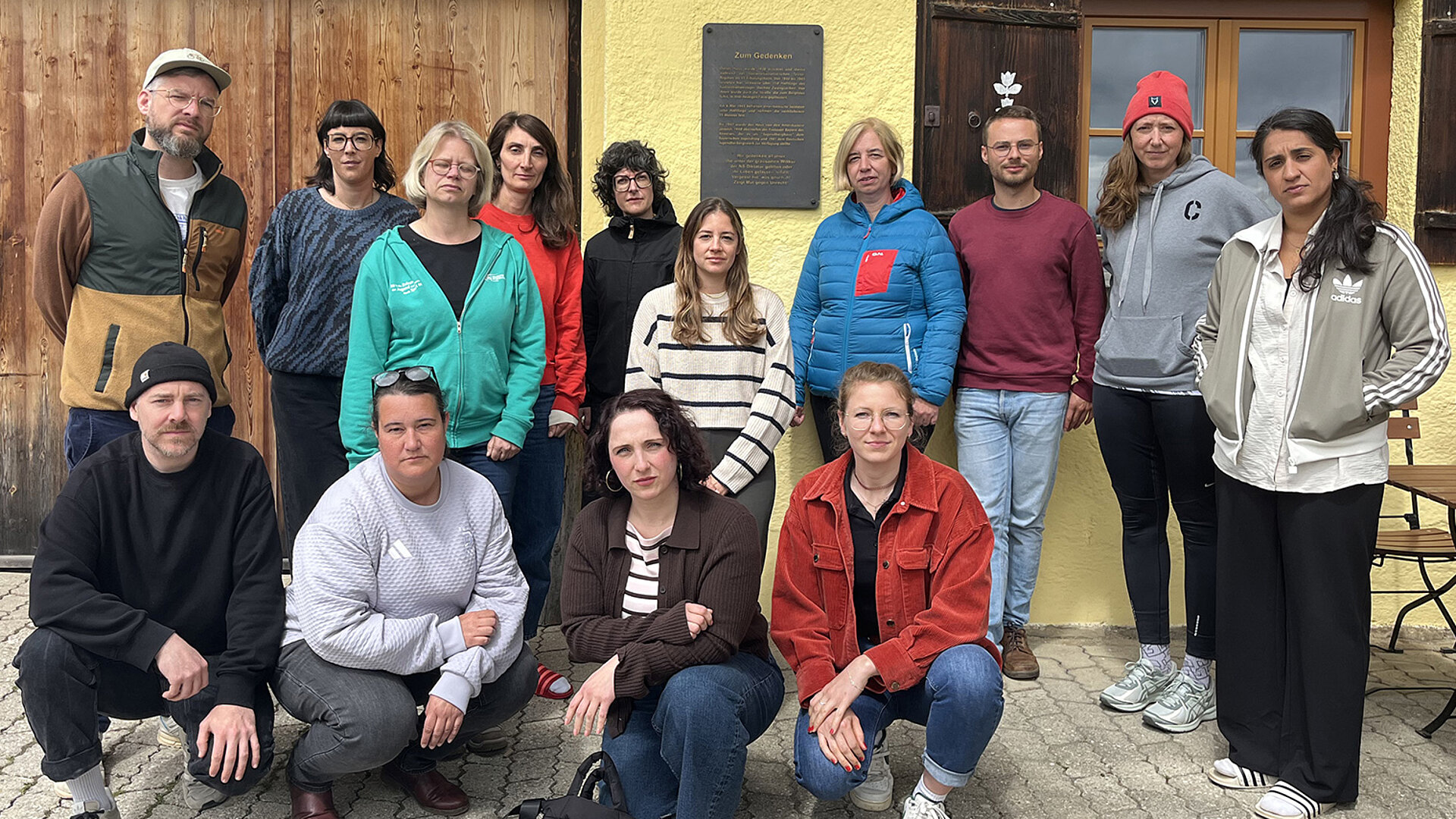 Gruppe von Teilnehmenden einer Erinnerungsfahrt der Deutschen Sportjugend steht und kniet vor einem Gebäude am Sudelfeld; im Hintergrund ist eine Gedenktafel zur Gründung angebracht.