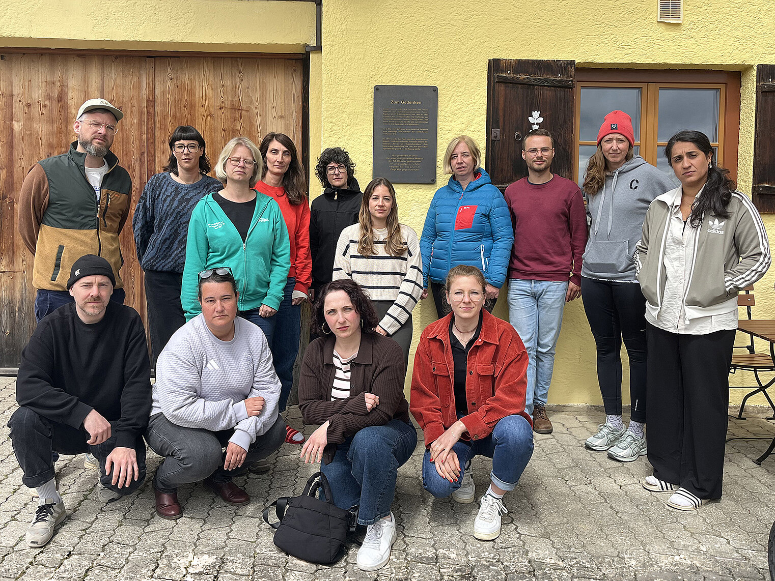 Gruppe von Teilnehmenden einer Erinnerungsfahrt der Deutschen Sportjugend steht und kniet vor einem Gebäude am Sudelfeld; im Hintergrund ist eine Gedenktafel zur Gründung angebracht.