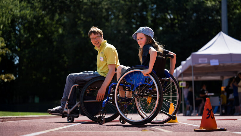 Ein Kind und ein junger Mensch in Sportrollstühlen auf einer Laufbahn bei einer Inklusionssportveranstaltung im Freien