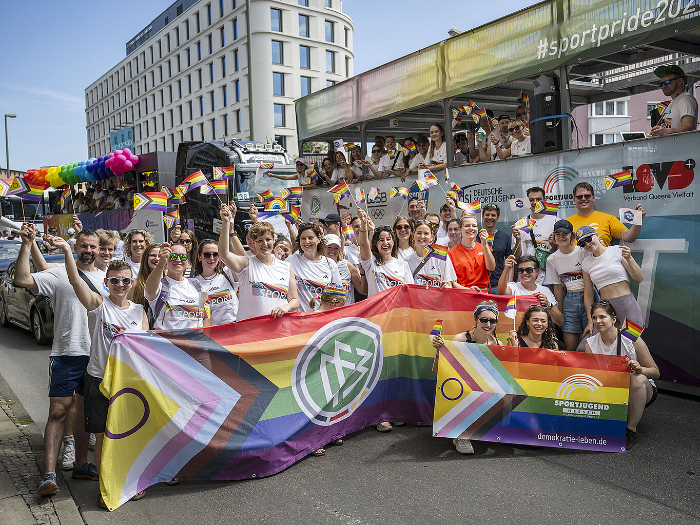 Gruppe von Teilnehmer*innen beim CSD Frankfurt mit Regenbogenflaggen und einem Banner. Sie tragen T-Shirts mit der Aufschrift „Loud and Proud im Sport“ und posieren vor einem bunt geschmückten Festwagen der Parade.