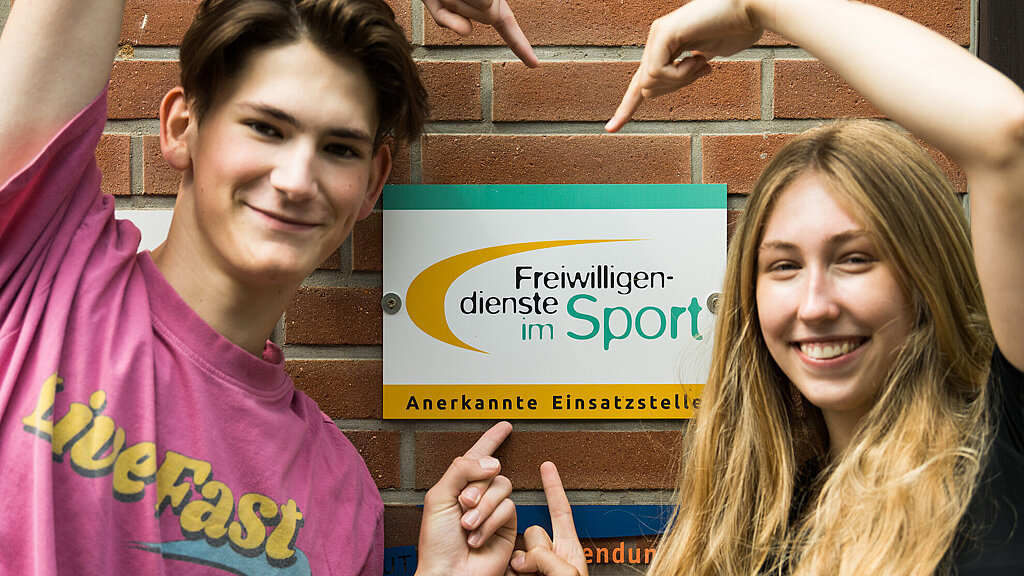 zwei Freiwilligendienstleistende umrahmen ein Schild "anerkannte Einsatzstelle"