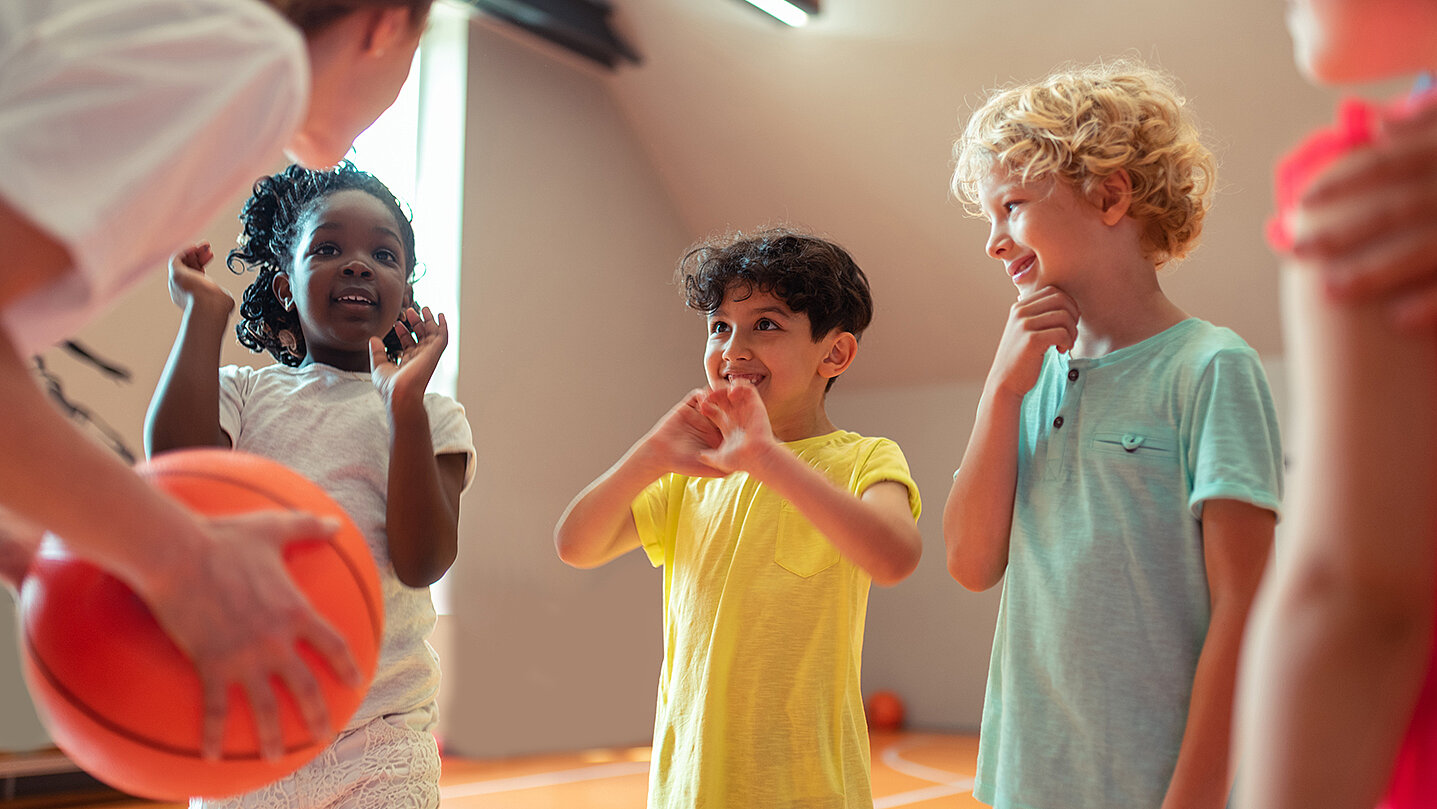 Eine Gruppe von vier Kindern steht in einer Sporthalle. Eine Trainerin hält einen Basketball, während die Kinder aufmerksam schauen und miteinander sprechen. Die Kinder zeigen verschiedene Emotionen, darunter Freude und Aufregung.