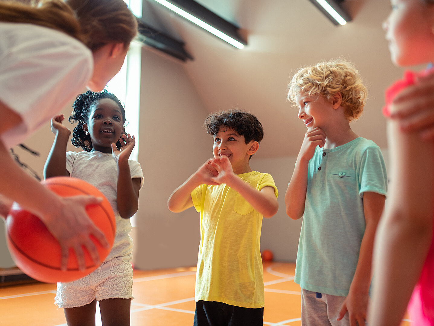 Eine Gruppe von vier Kindern steht in einer Sporthalle. Eine Trainerin hält einen Basketball, während die Kinder aufmerksam schauen und miteinander sprechen. Die Kinder zeigen verschiedene Emotionen, darunter Freude und Aufregung.