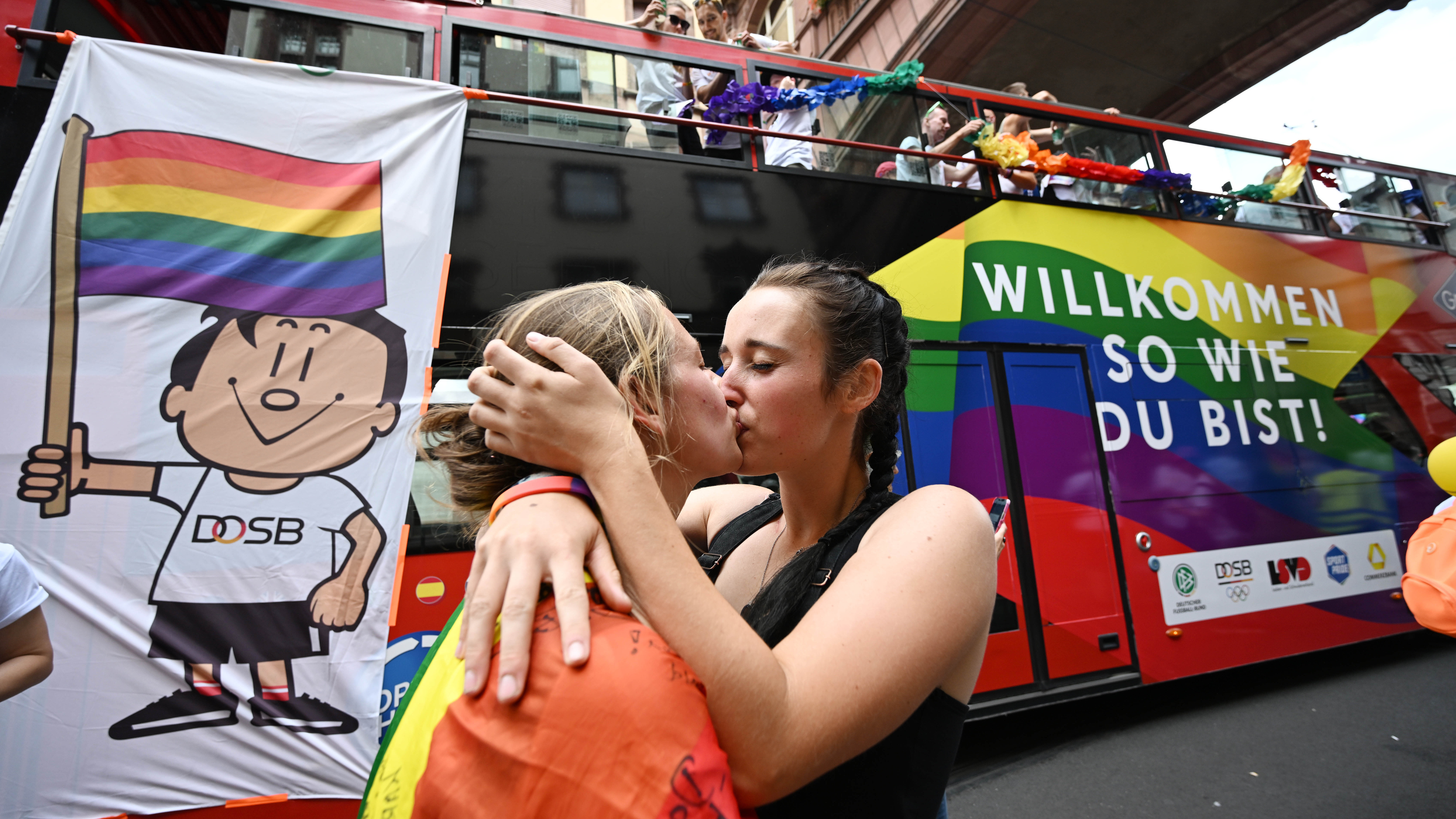 Zwei Frauen küssen sich während der Pride Parade vor einem bunten Bus mit der Aufschrift 'Willkommen so wie du bist!' 