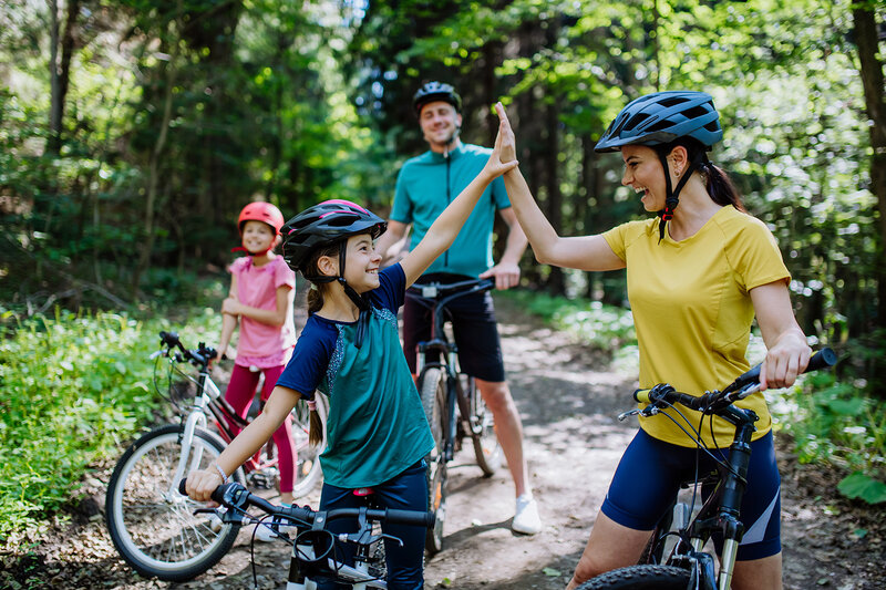 Vier Radfahrer*innen, ein Kind und eine Frau, geben sich im Wald einen High-Five, während zwei weitere Personen mit Fahrrädern im Hintergrund stehen.