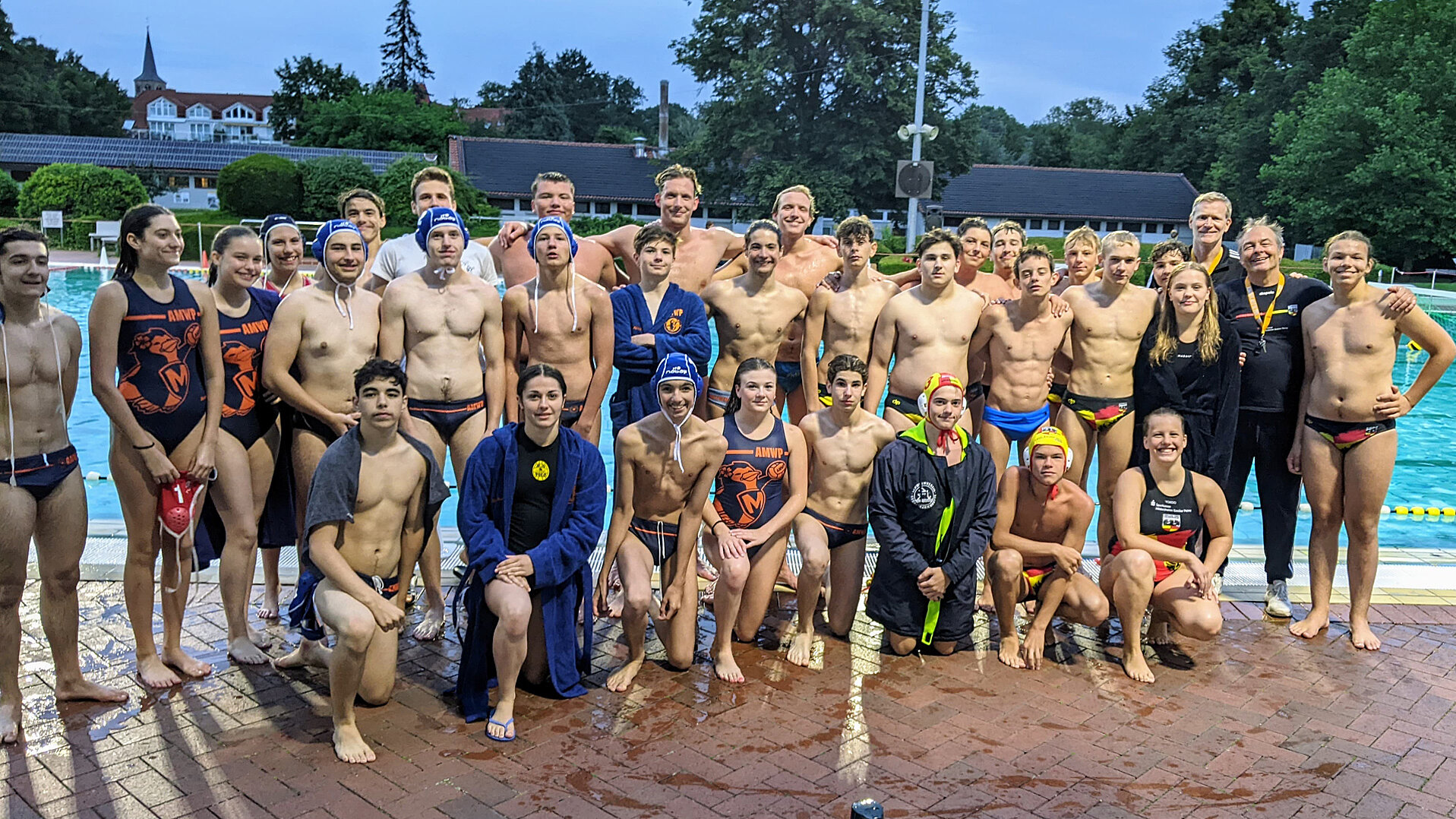 Eine Gruppe von Jugendlichen und jungen Erwachsenen steht am Beckenrand eines Schwimmbades. Sie tragen Schwimmbekleidung und einige haben Handtücher um die Schultern. Im Hintergrund ist das Schwimmbecken und Bäume zu sehen. Die Stimmung wirkt freundlich und sportlich.