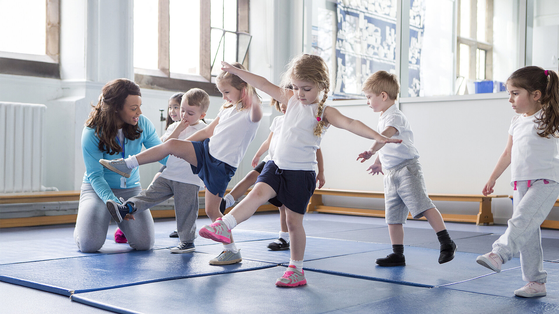Fünf Kinder und eine Betreuerin üben gemeinsam Bewegungsübungen auf blauen Matten in einem hellen Raum, gefördert durch Proficlubs für mehr Bewegung in Kitas und Schulen.