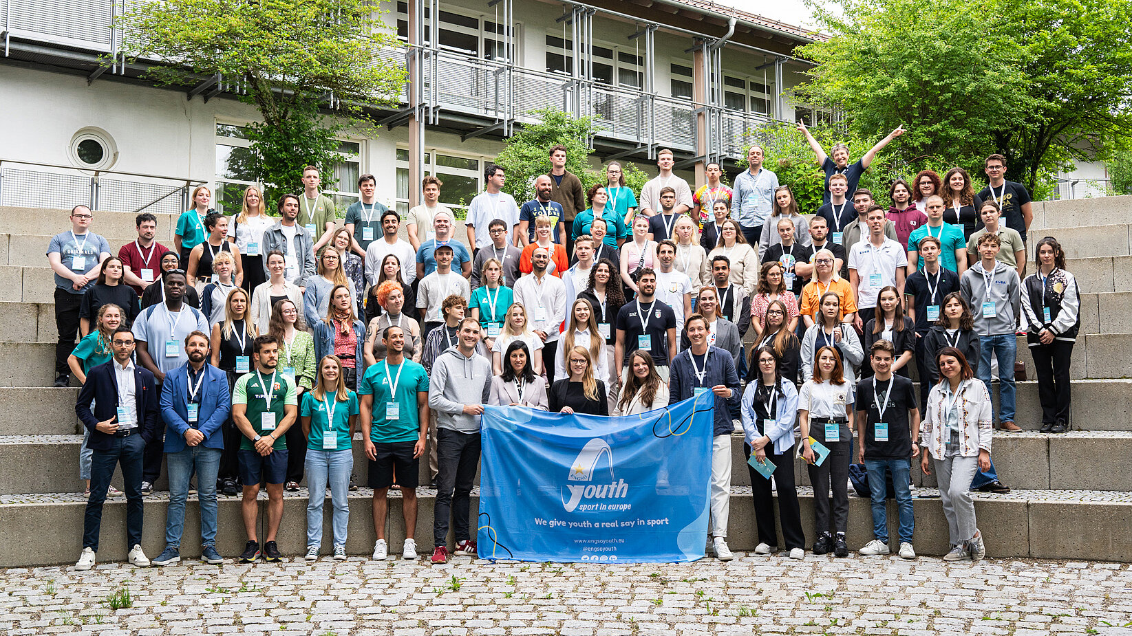 Gruppenfoto von etwa 80 Jugendlichen und jungen Erwachsenen vor einem Gebäude in München bei der 2. Ausgabe der Europäischen Plattform für Jugend und Sport von ENGSO Youth, einige halten ein blaues Banner mit dem ENGSO Youth Logo und dem Motto 'The young people's voice in sport'