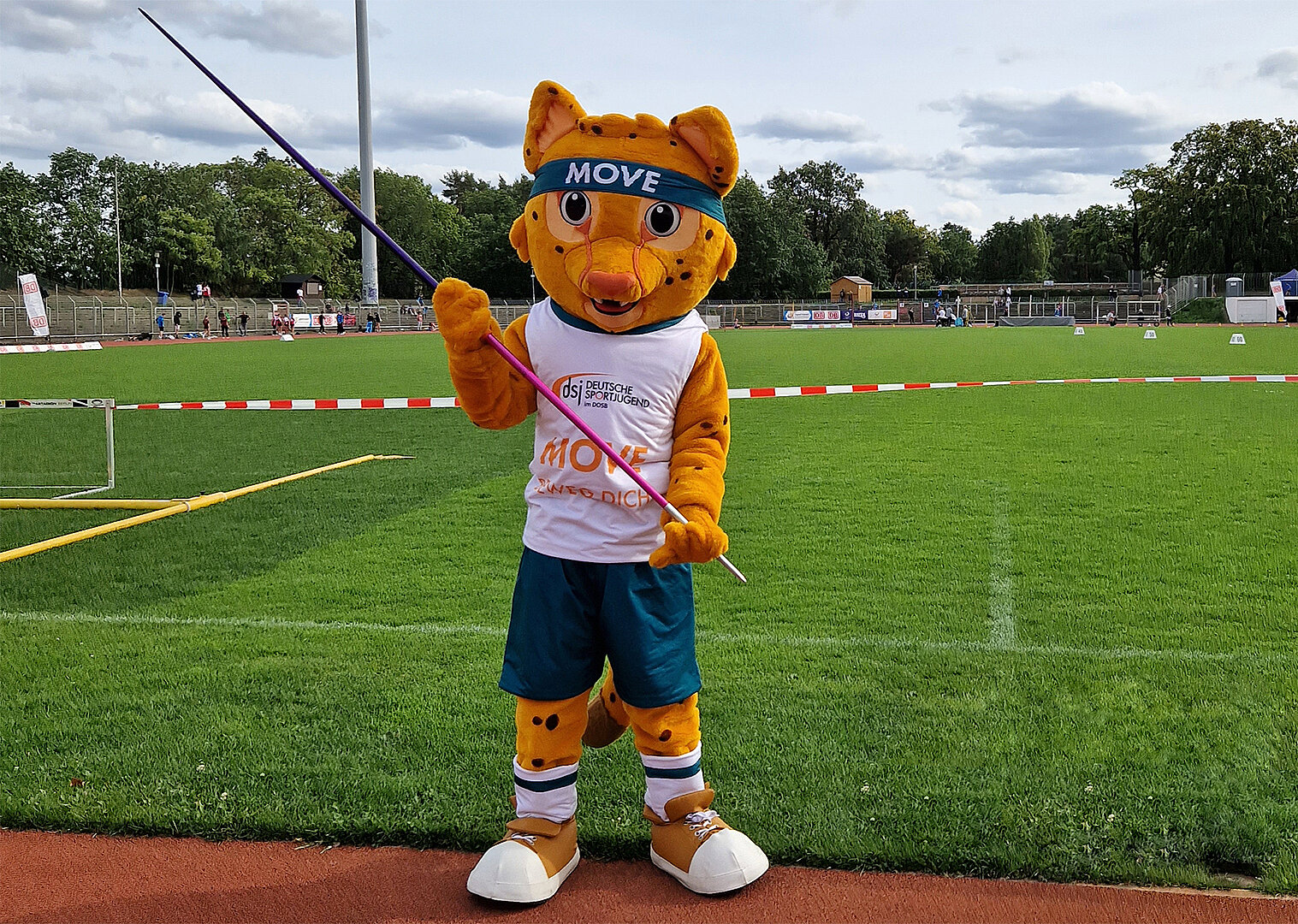 Maskottchen Movelino, ein orange-gelber Gepard mit Stirnband 'MOVE', trägt ein weißes Trikot mit 'Move' und steht auf einer Sportanlage mit Speerwurfgerät