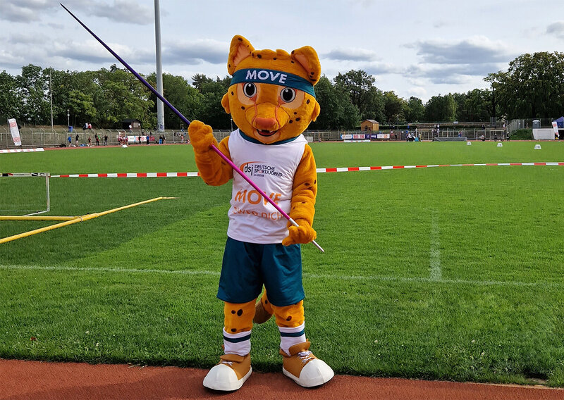 Maskottchen Movelino, ein orange-gelber Gepard mit Stirnband 'MOVE', trägt ein weißes Trikot mit 'Move' und steht auf einer Sportanlage mit Speerwurfgerät