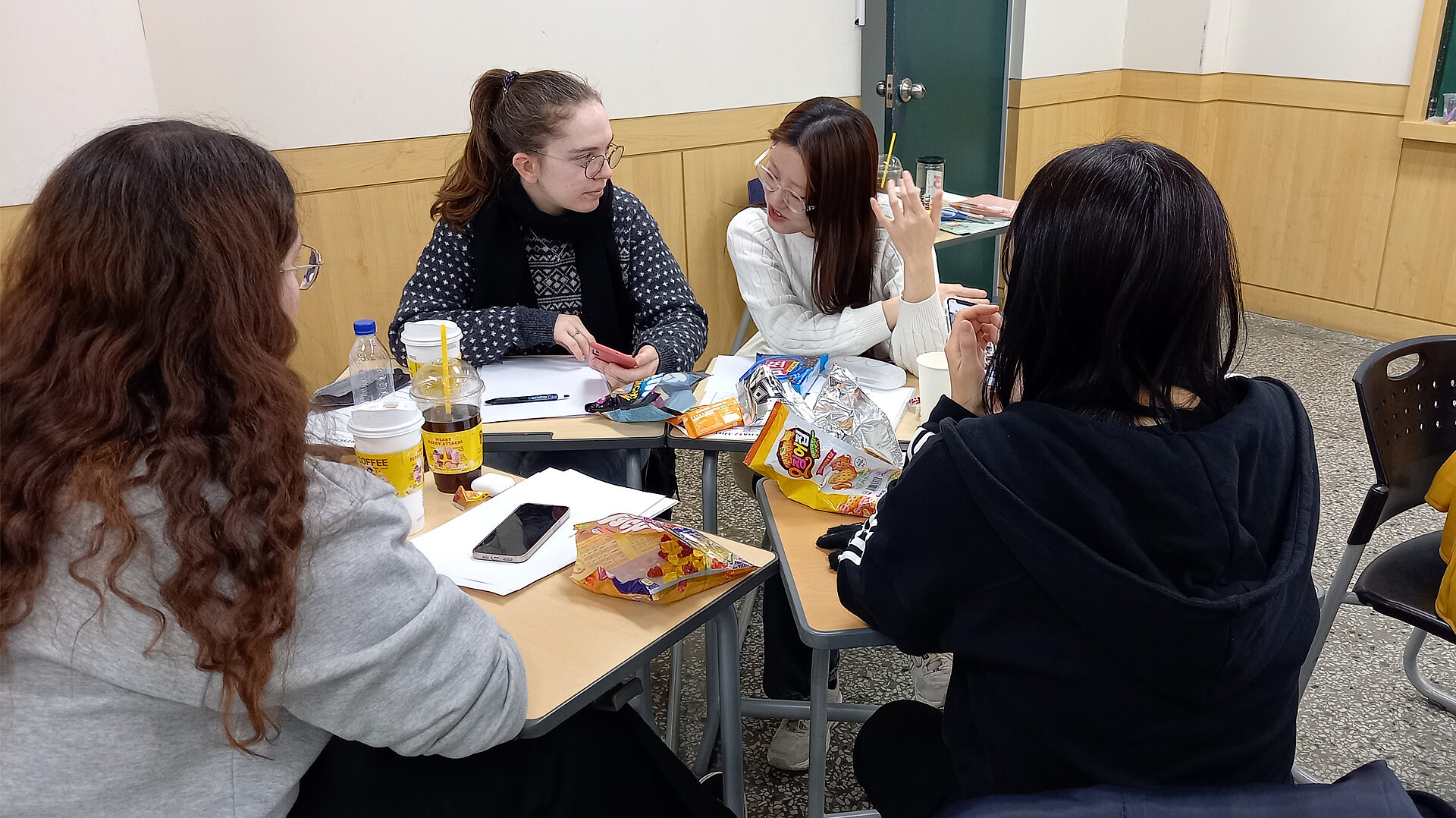 Vier Personen sitzen an einem Tisch, umgeben von Snacks und Getränken. Sie diskutieren miteinander, während einige Notizen und ein Smartphone auf dem Tisch liegen. Der Raum hat helle Wände und einen grünen Türrahmen.