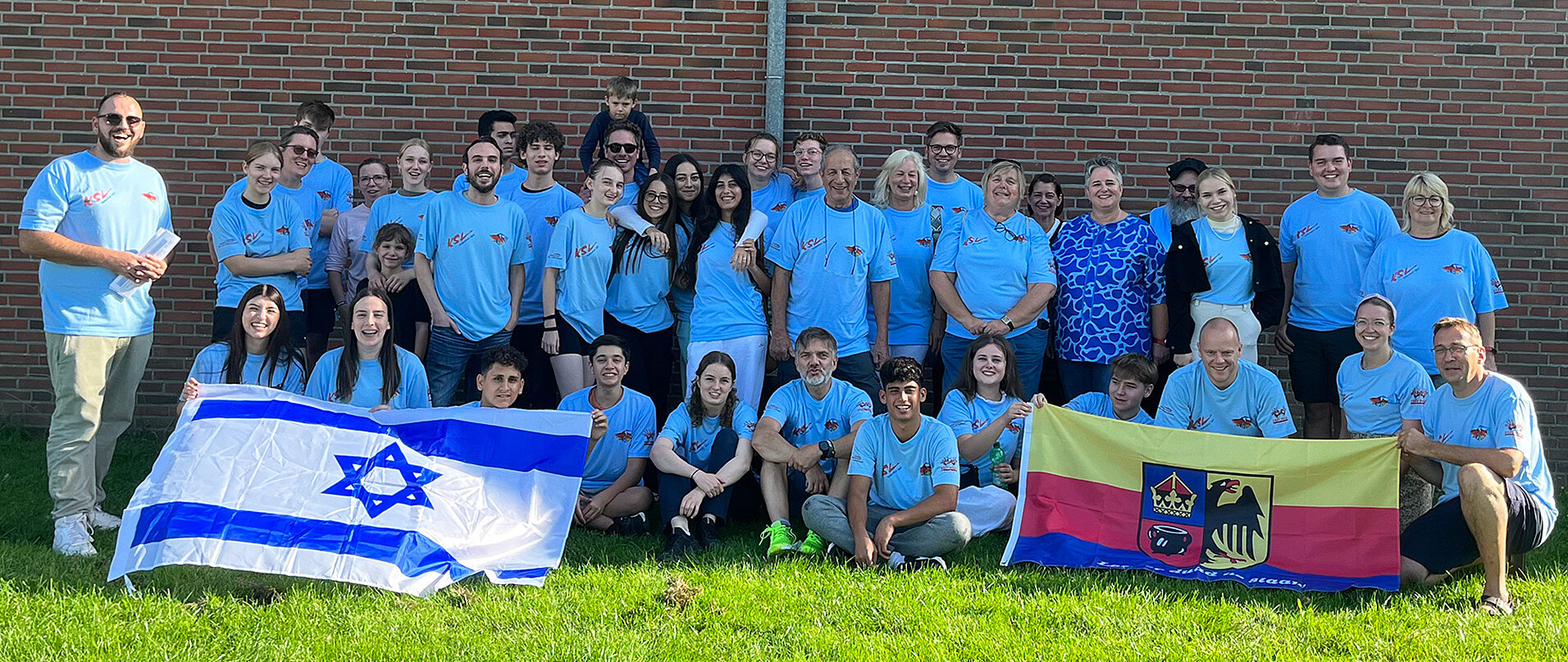 Eine Gruppe von etwa 30 Personen in hellblauen T-Shirts steht auf einer grünen Wiese vor einer Ziegelwand. Einige halten Flaggen, darunter eine israelische Flagge und eine Flagge mit einem Wappen. Die Stimmung wirkt freundlich und gemeinschaftlich.