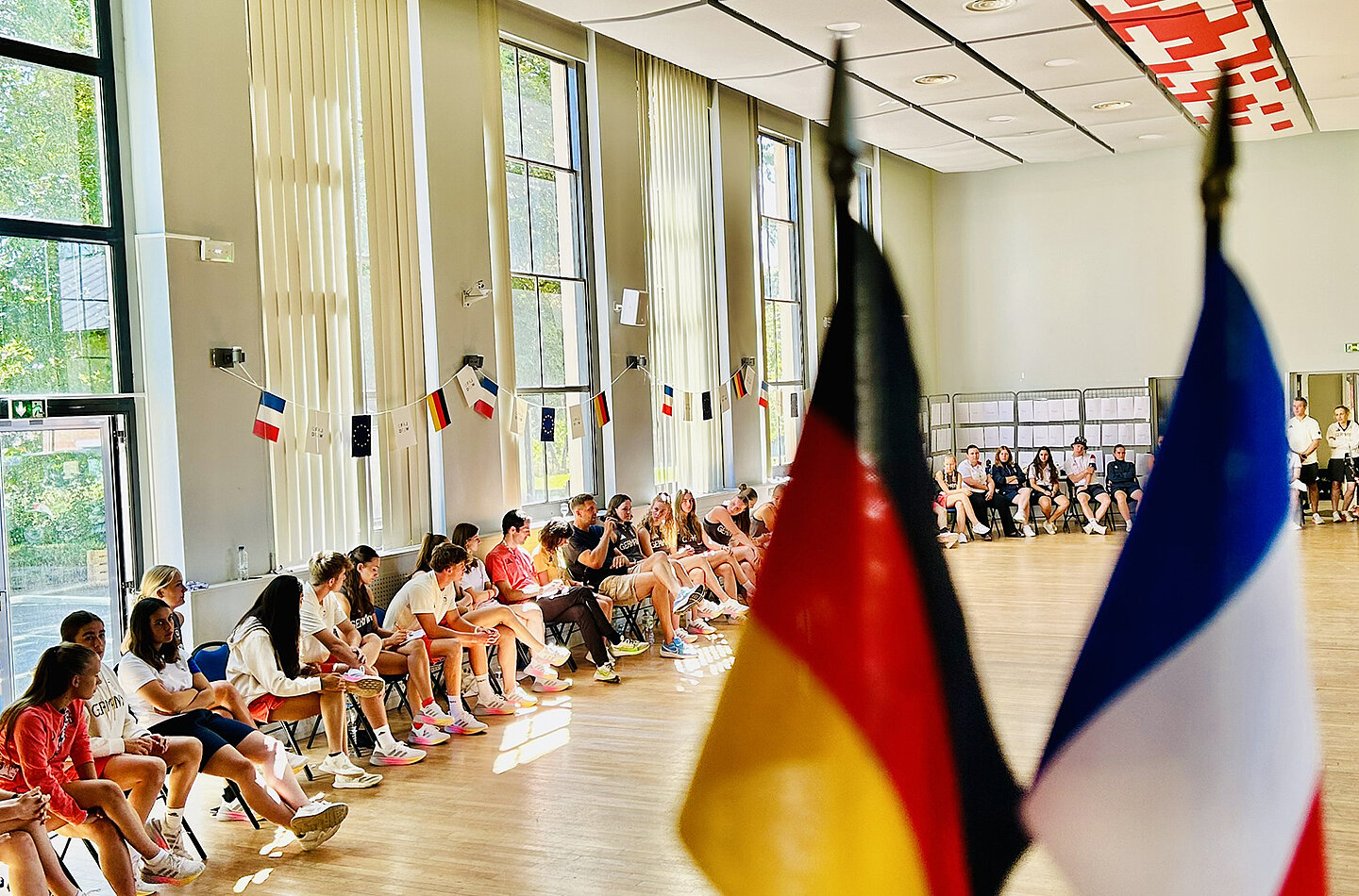 Im Vordergrund sind die Flaggen von Deutschland und Frankreich zu sehen. Im Hintergrund sitzen zahlreiche Jugendliche in einem hellen Raum mit großen Fenstern. An den Wänden hängen Flaggen verschiedener Länder. Es herrscht eine freundliche, gesellige Atmosphäre.