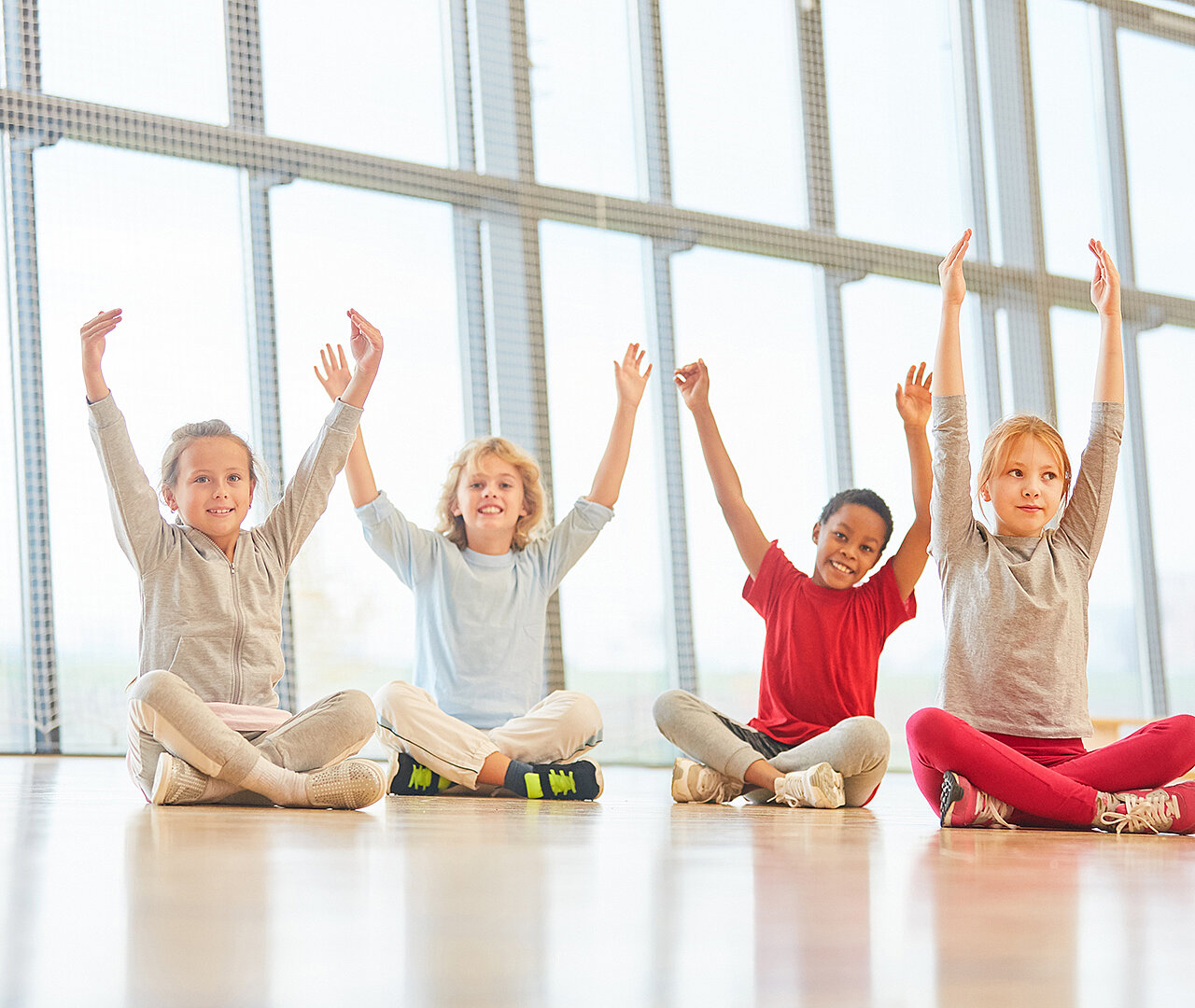 Vier Kinder sitzen auf dem Boden in einem hellen Raum mit großen Fenstern. Sie haben die Arme erhoben und lächeln. Die Kinder tragen bequeme Kleidung in verschiedenen Farben und scheinen Freude zu haben.