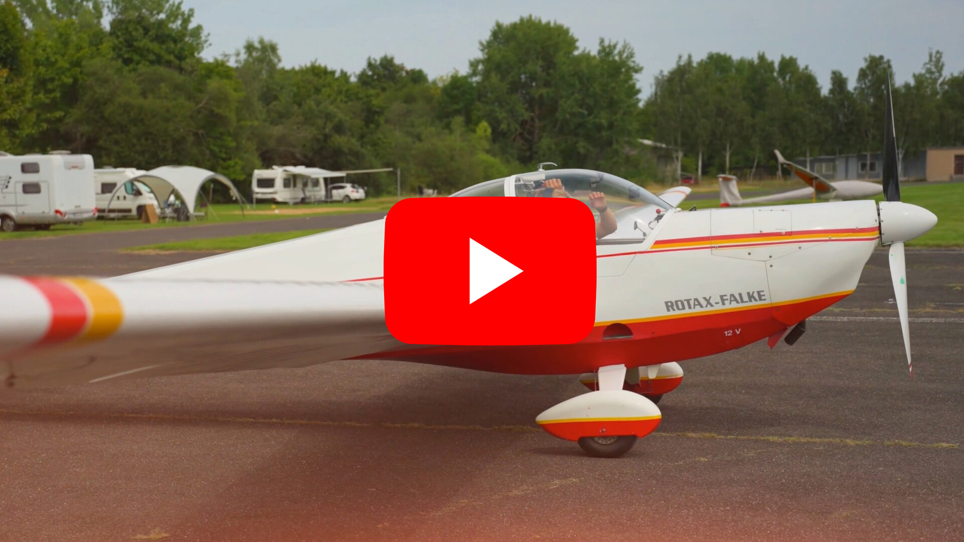Ein einmotoriges Sportflugzeug mit weißem Rumpf und orangeroten Akzenten steht auf einem Flugplatz. Auf der Seite des Flugzeugs ist die Aufschrift 'ROTAX-FALKE' zu sehen. Im Hintergrund sind mehrere Wohnwagen und Zelte auf einer Wiese vor einer Baumreihe zu erkennen. Der Himmel ist bewölkt, und das Flugzeug steht auf einer asphaltierten Fläche.