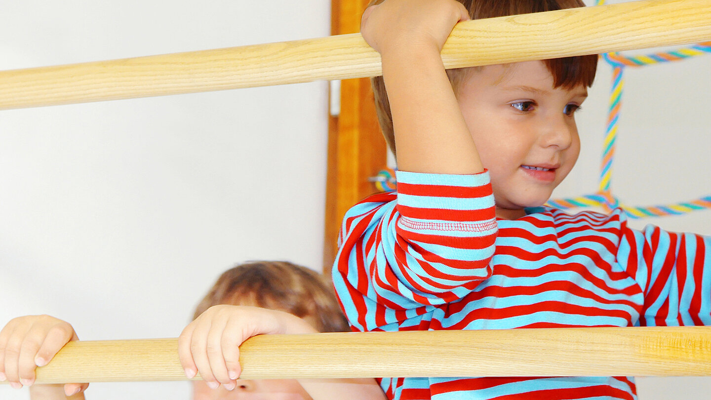 Zwei Kinder spielen in einem Raum. Ein Junge mit gestreiftem Oberteil hält sich an einem Holzklettergerüst fest und lächelt. Im Hintergrund ist ein weiteres Kind zu sehen, das ebenfalls am Gestell greift.