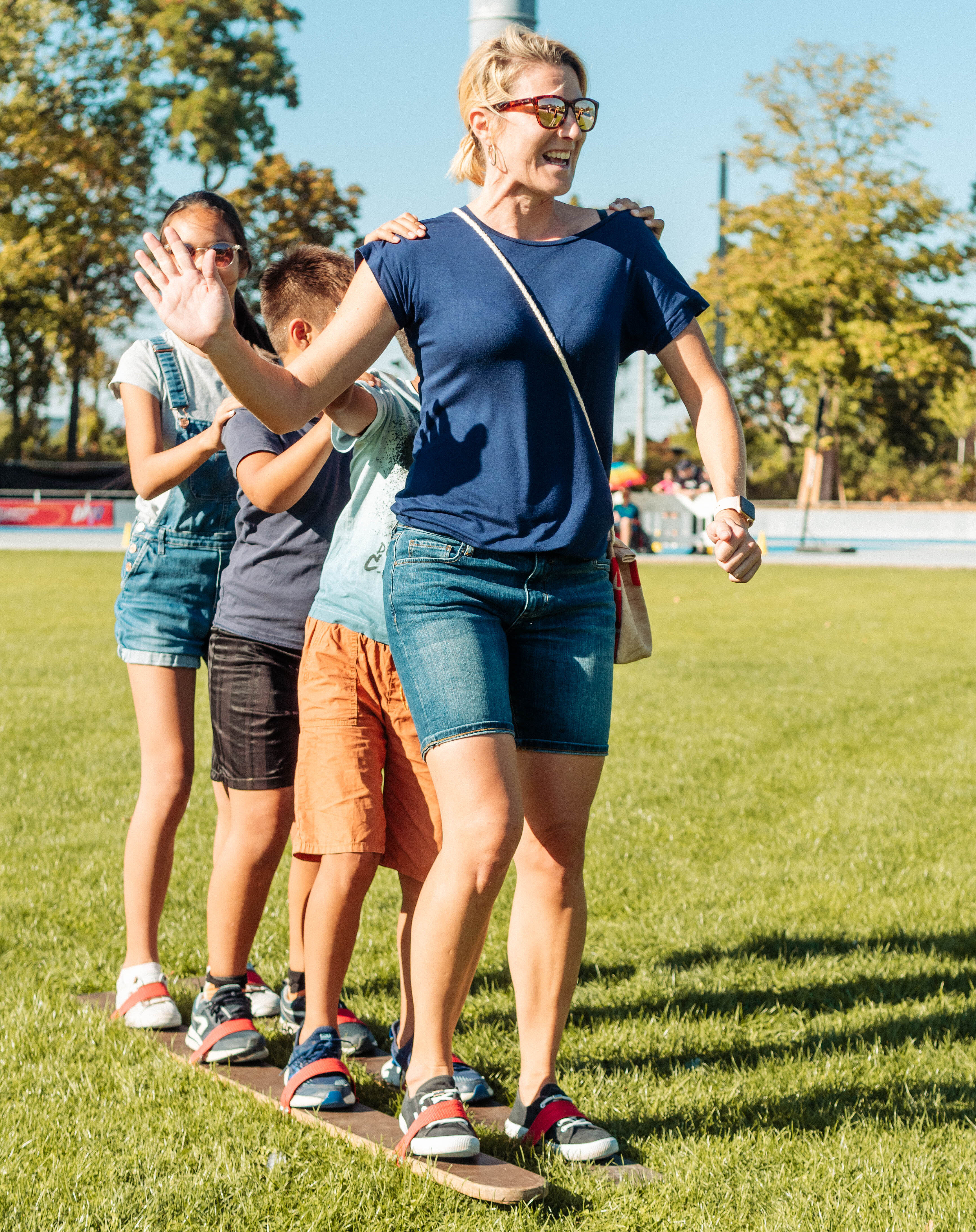 Eine Gruppe von vier Personen steht auf einem schmalen Brett im Freien. Eine Frau führt die Gruppe an, während die anderen Kinder hinter ihr stehen und sich an ihr festhalten. Alle lachen und haben Spaß. Im Hintergrund sind Bäume und eine Wiese zu sehen.