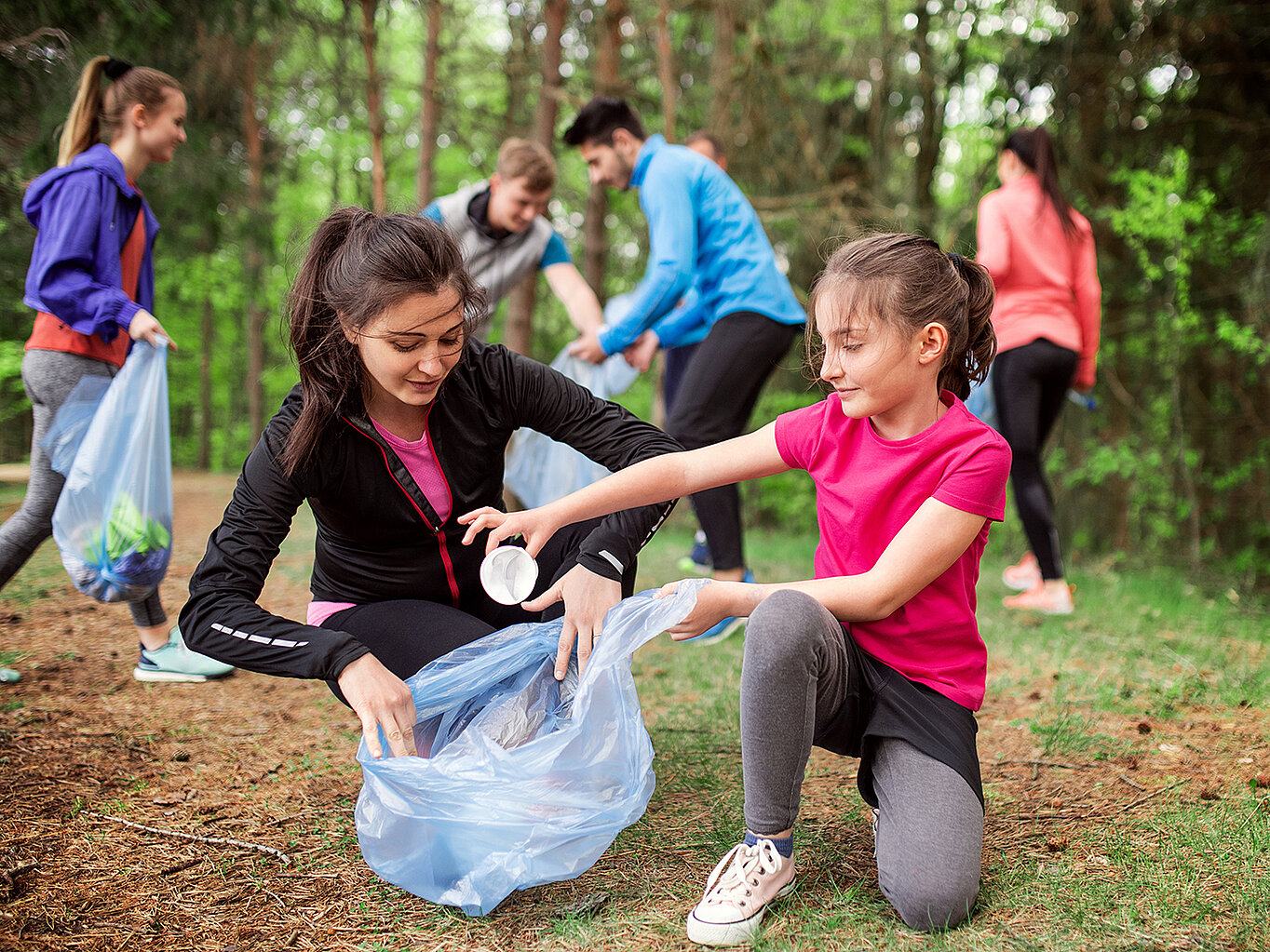 Eine Gruppe von Menschen sammelt Müll im Wald. Zwei Mädchen, eines in schwarzer und das andere in pinker Sportkleidung, arbeiten zusammen, um Abfall in einen blauen Müllsack zu legen. Im Hintergrund sind weitere Personen zu sehen, die ebenfalls Müll einsammeln.