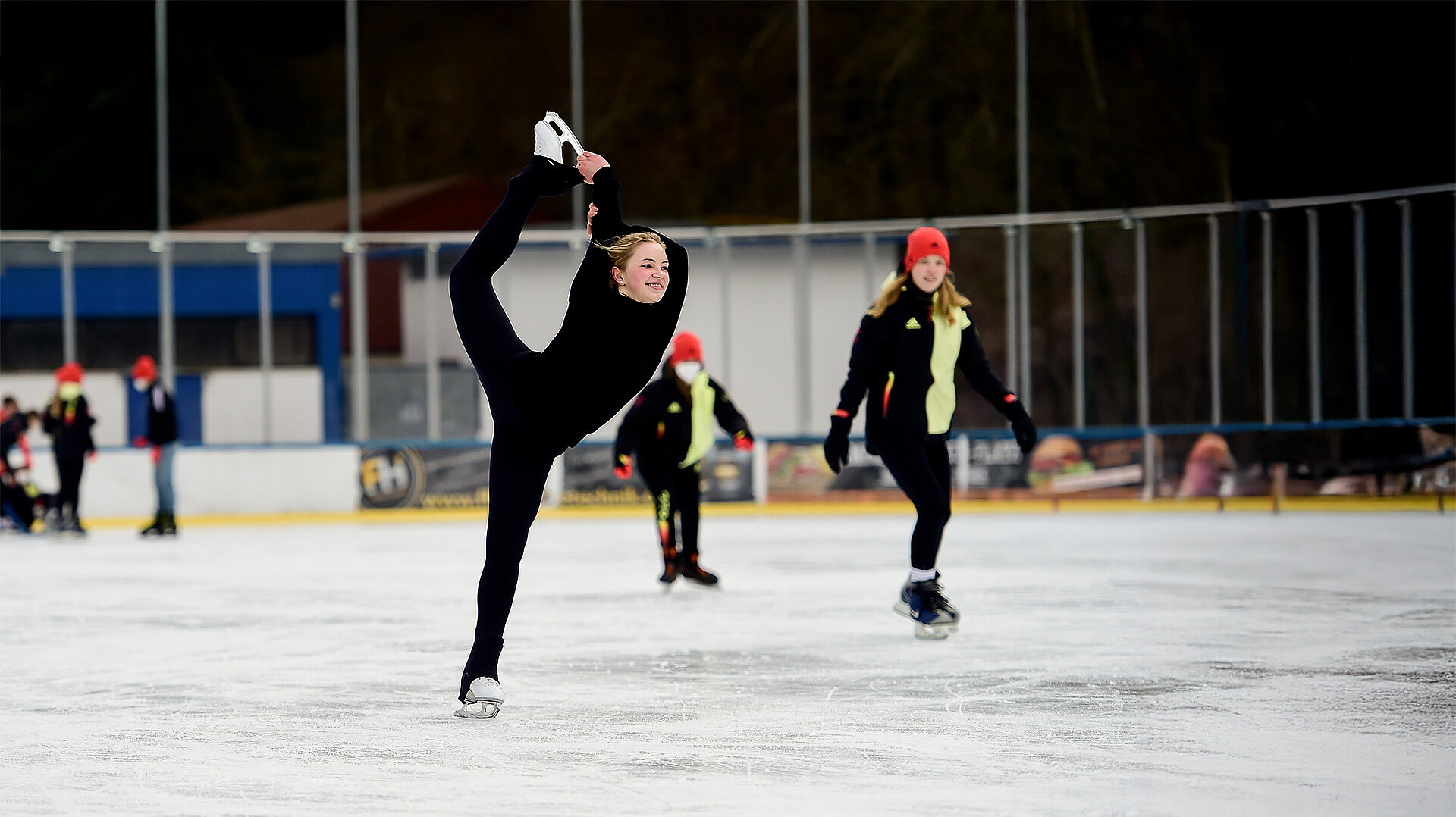 Jugendliche beim Eiskunstlauf auf einer Eisbahn, eine Läuferin führt eine Dehnung mit einem Bein in der Hand aus, andere tragen rote Mützen, Bewerbung für das Olympische Jugendlager Mailand Cortina 2026