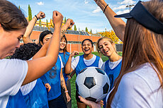 Eine Gruppe von Frauen in blauen Trikots feiert auf einem Fußballfeld. Sie heben die Arme und zeigen Freude, während eine Spielerin einen Fußball in der Hand hält. Der Hintergrund zeigt einen klaren Himmel und Bäume.