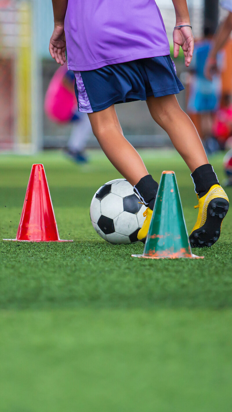 Kind in lila Shirt und blauen Shorts dribbelt Fußball zwischen roten und grünen Hütchen auf Sportplatz