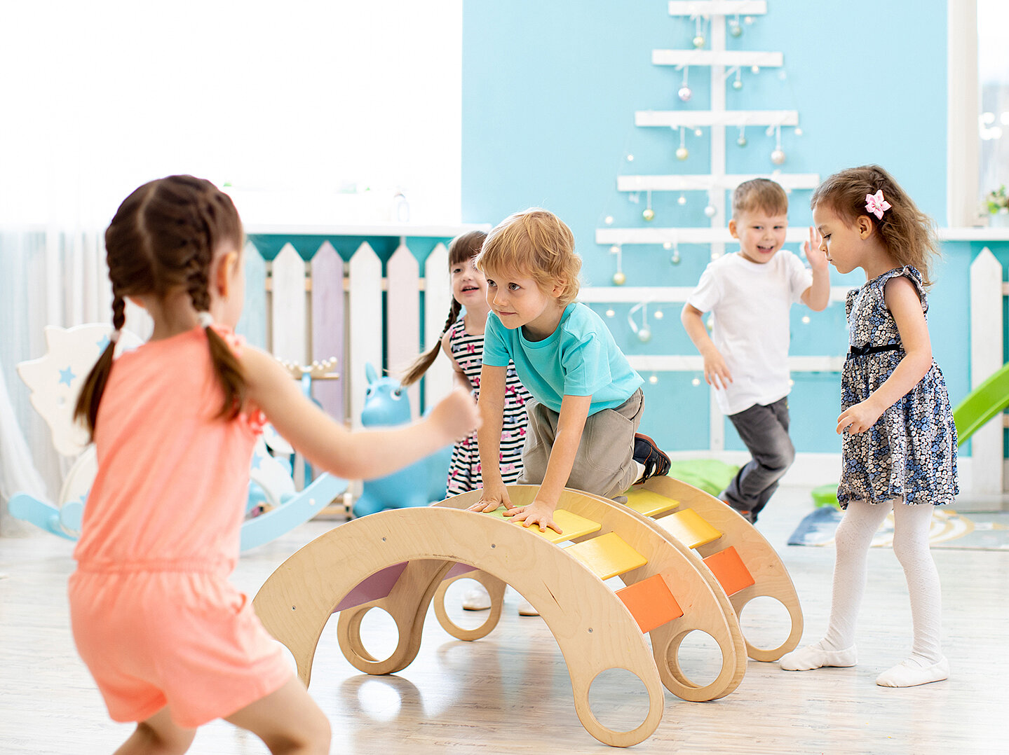 Mehrere Kinder spielen und bewegen sich gemeinsam in einem hellen Bewegungsraum, klettern auf bunten Holz‑Spielgeräten und fördern dabei Gleichgewicht, Koordination und Freude an Bewegung.